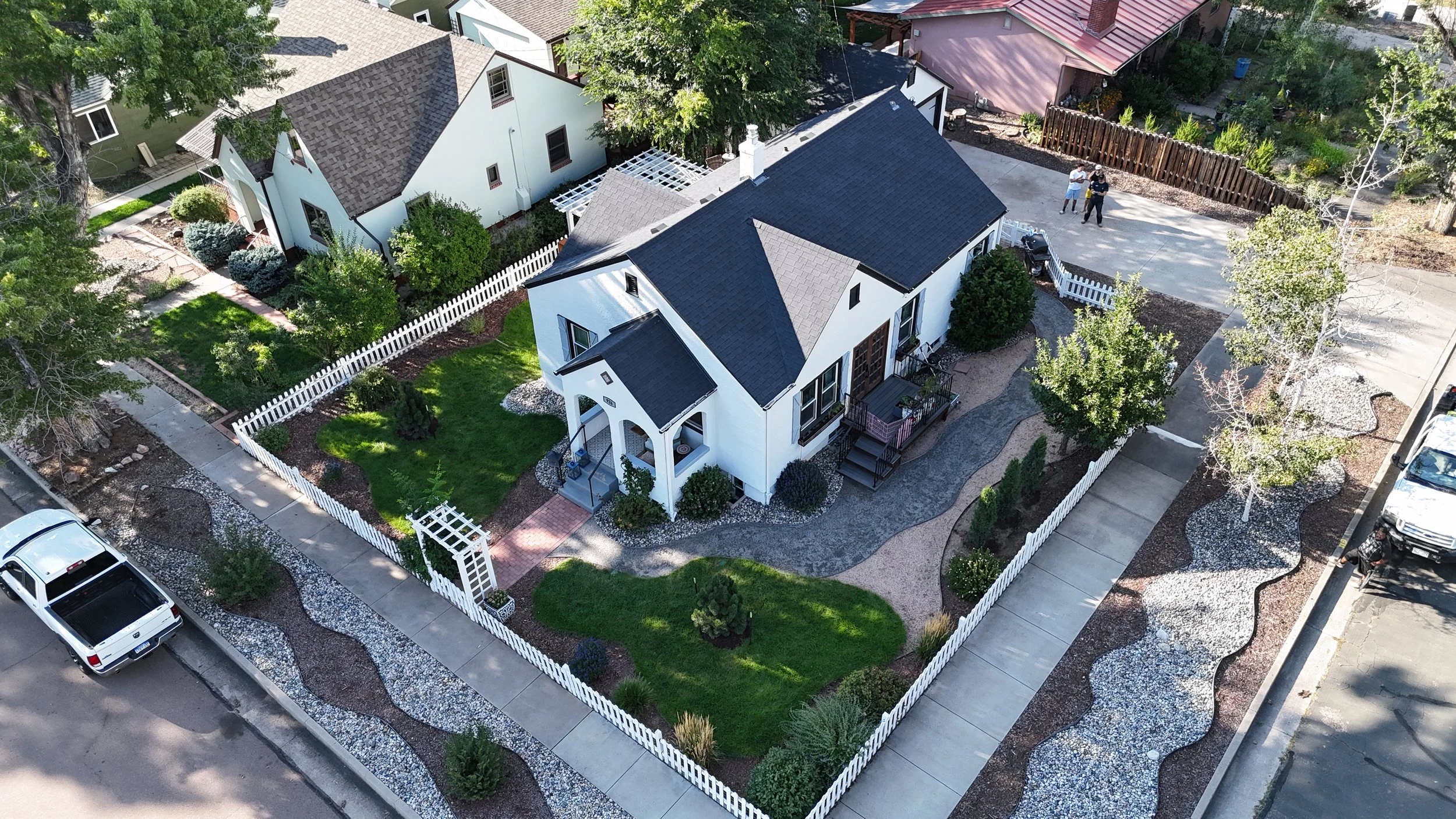 Aerial view of a white house with a dark roof, surrounded by a white picket fence, green lawn, and landscaped garden with trees and bushes. There are neighboring houses and parked cars visible along the street.