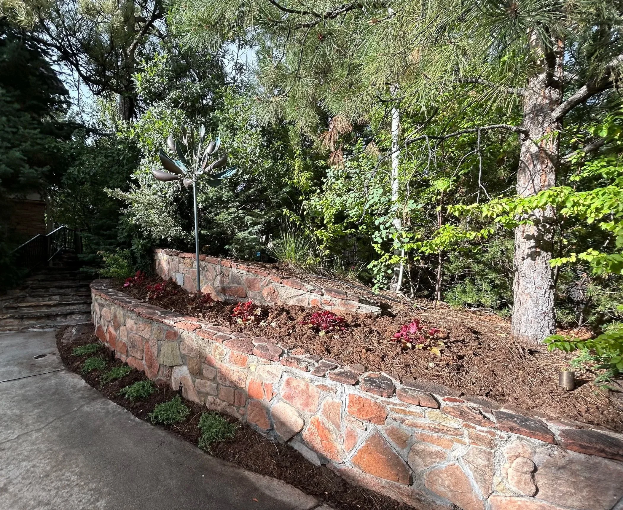 A stone retaining wall with a flower bed on top, containing plants and small flowers, adjacent to a concrete walkway with stairs leading up to a garden area, surrounded by trees and green foliage.