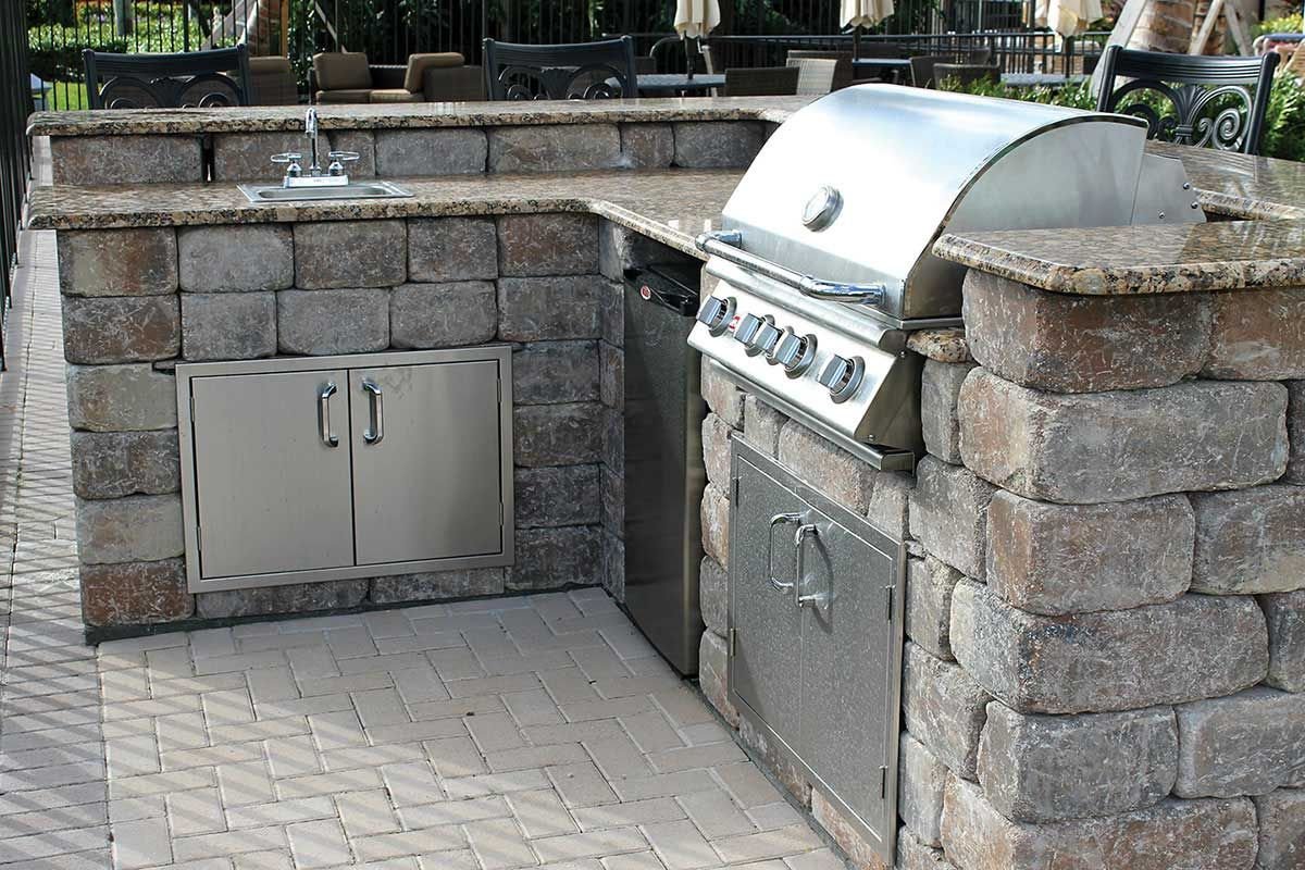 Outdoor built-in kitchen with stone brick structure, stainless steel grill, small sink, and storage cabinets, set on a paver patio.