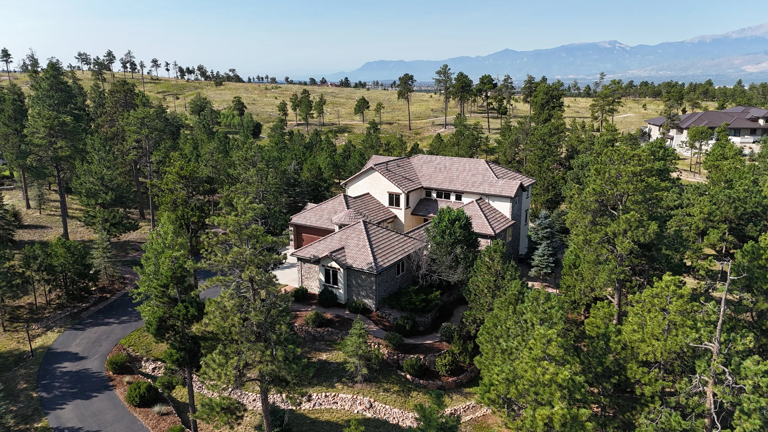 A large house with a tiled roof is surrounded by tall pine trees in a hilly landscape, with distant mountains in the background.