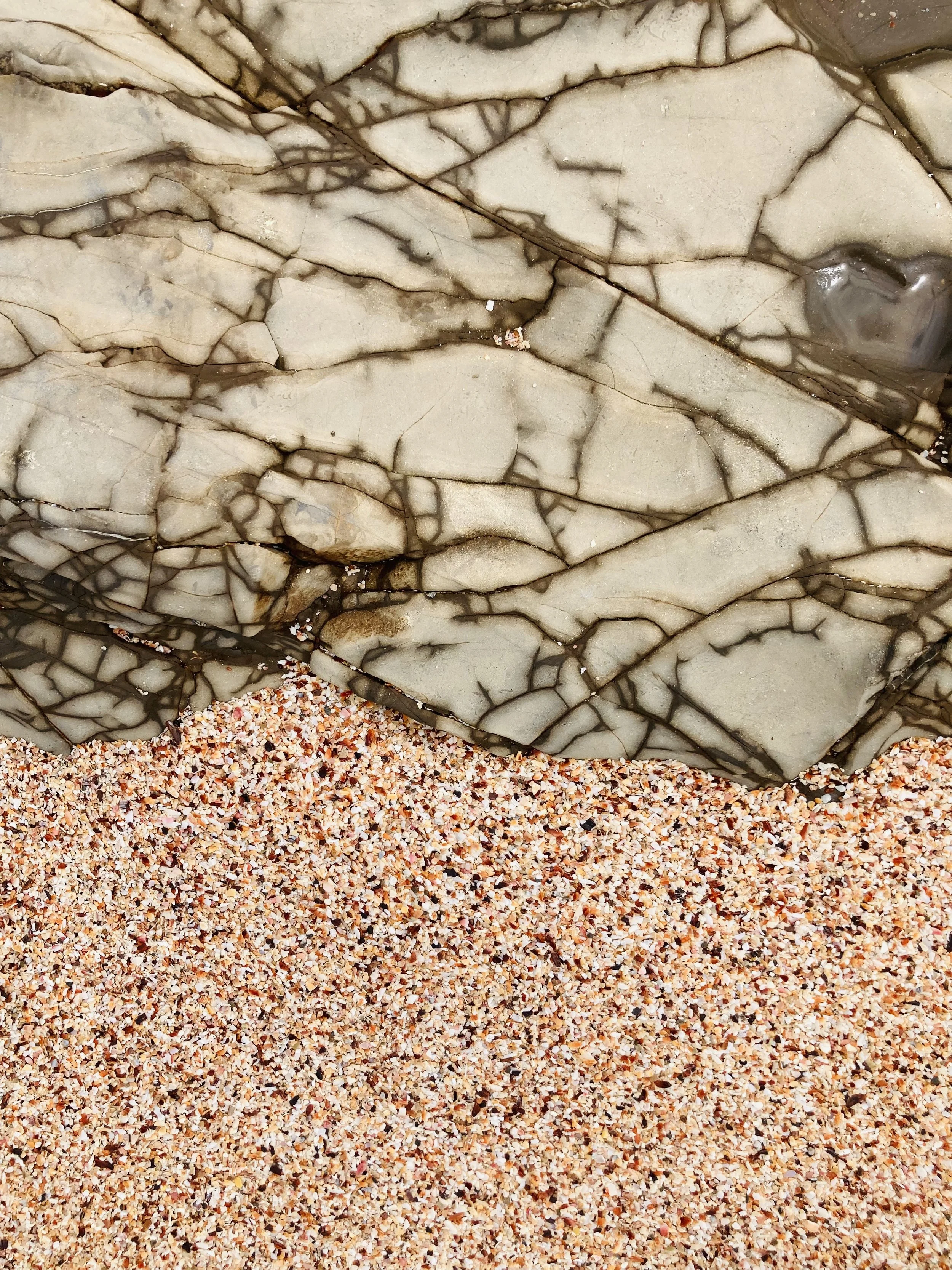 Close-up photo of a large rock with a pattern of dark veins, lying partly on multicolored sand.