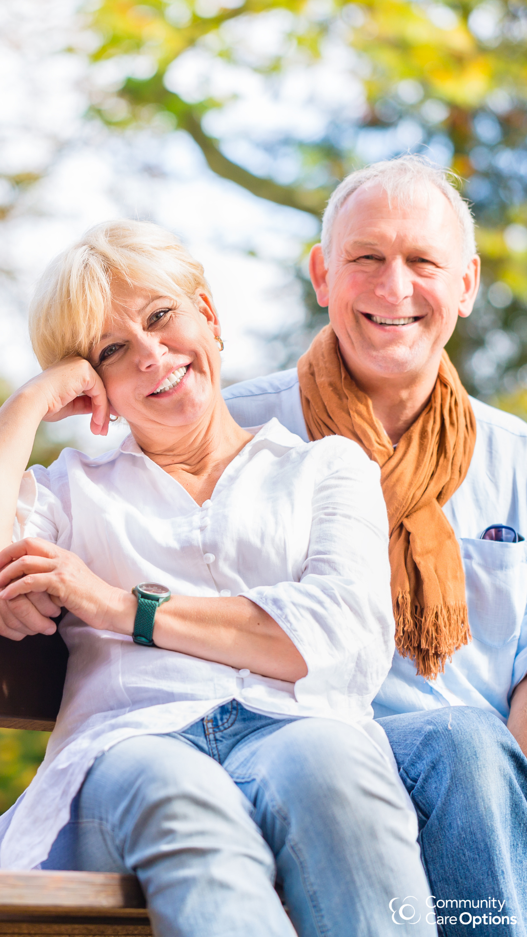 Smiling elderly woman and man sitting outdoors on a bench, surrounded by trees.