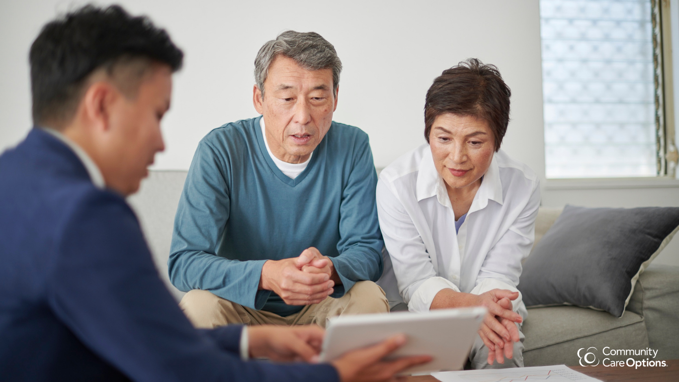 Two elderly adults, a man and a woman, sitting on a sofa and looking at a tablet held by a younger caregiver, who is dressed in a suit, during a consultation in a bright room.