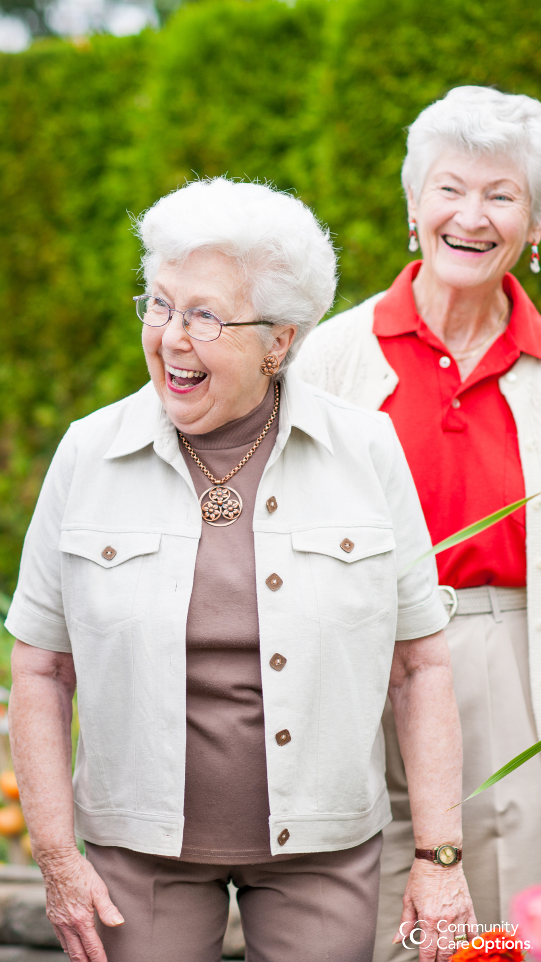 Two elderly women smiling and laughing outdoors with a green hedge in the background. They are dressed casually, one wearing a beige vest and glasses, the other in a red polo shirt and white cardigan.