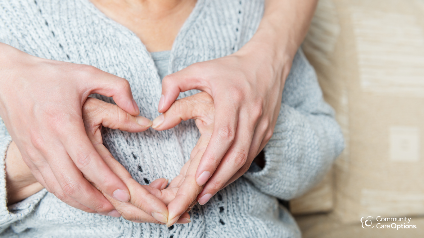 Multiple hands forming a heart shape over a person's chest, wearing a gray knitted sweater.