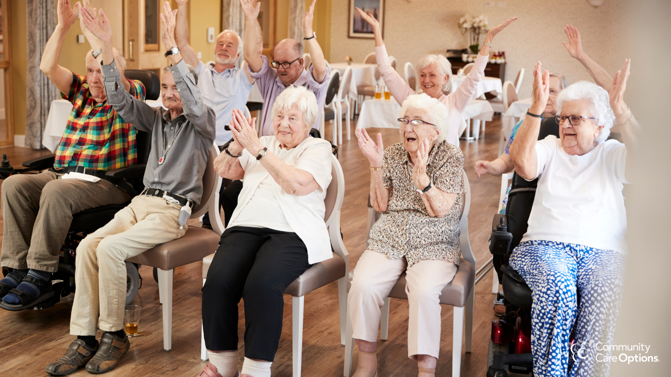Group of elderly people sitting and engaging in an activity, with some raising their hands in a cheerful and lively setting, possibly a community center or care facility.