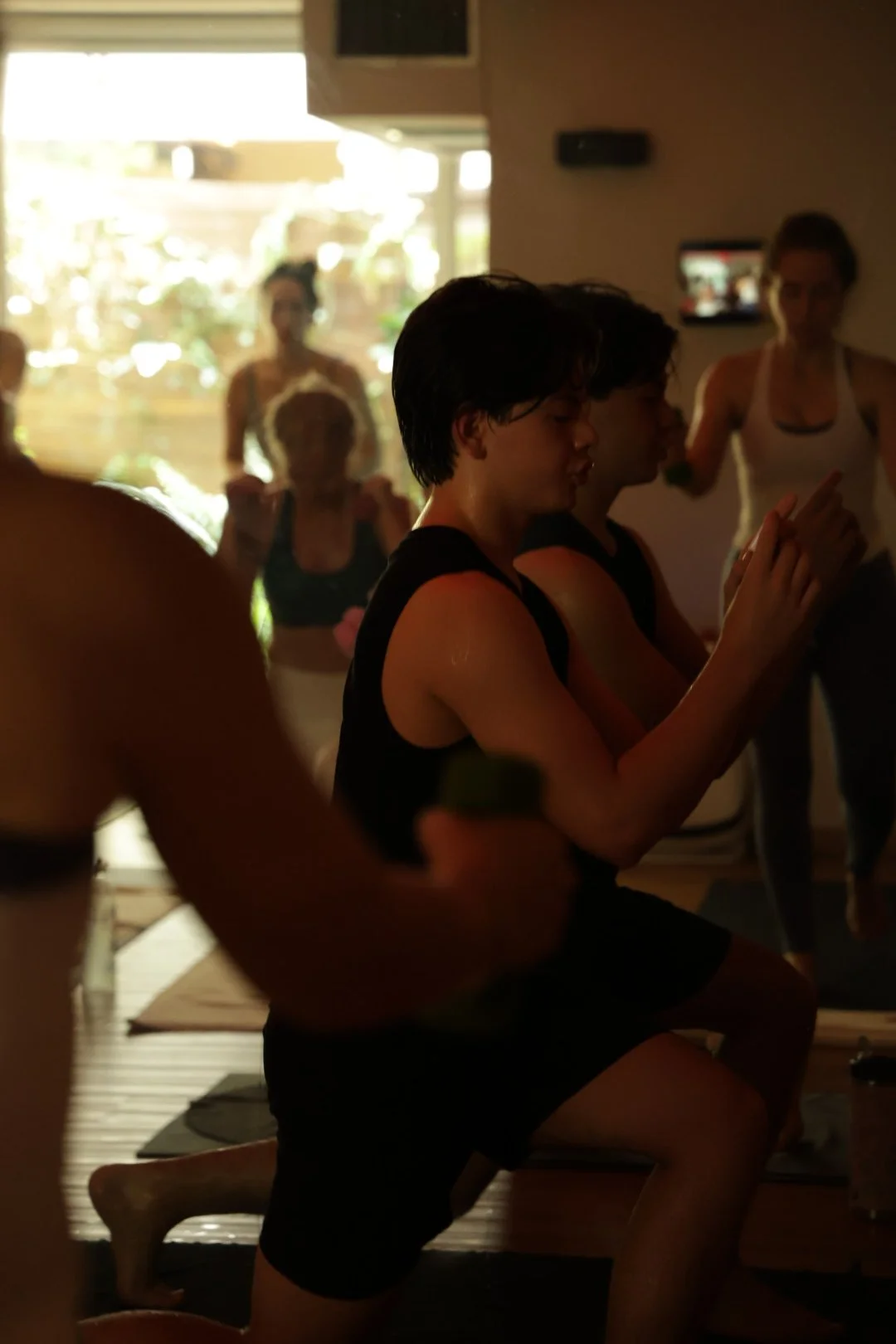 Grupo de personas participando en una clase de yoga en interior, en posición de cuclillas con los ojos cerrados.