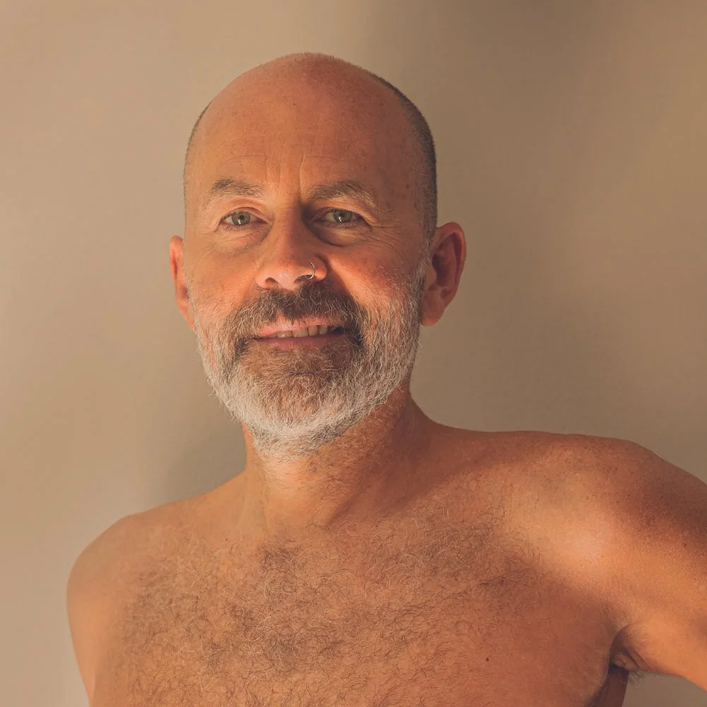 Hombre calvo de cabello gris y barba, tomando una selfie sin camisa, sonriendo frente a un fondo neutro.