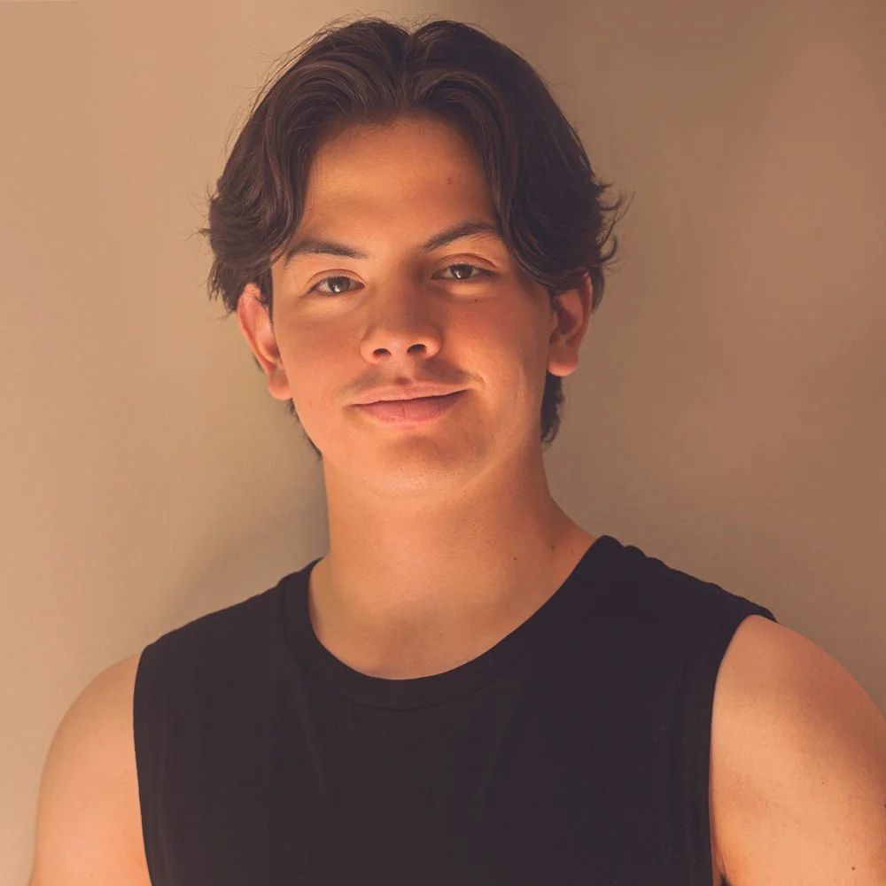 Joven con cabello castaño, sonrisa leve, viste camiseta sin mangas, fondo beige.