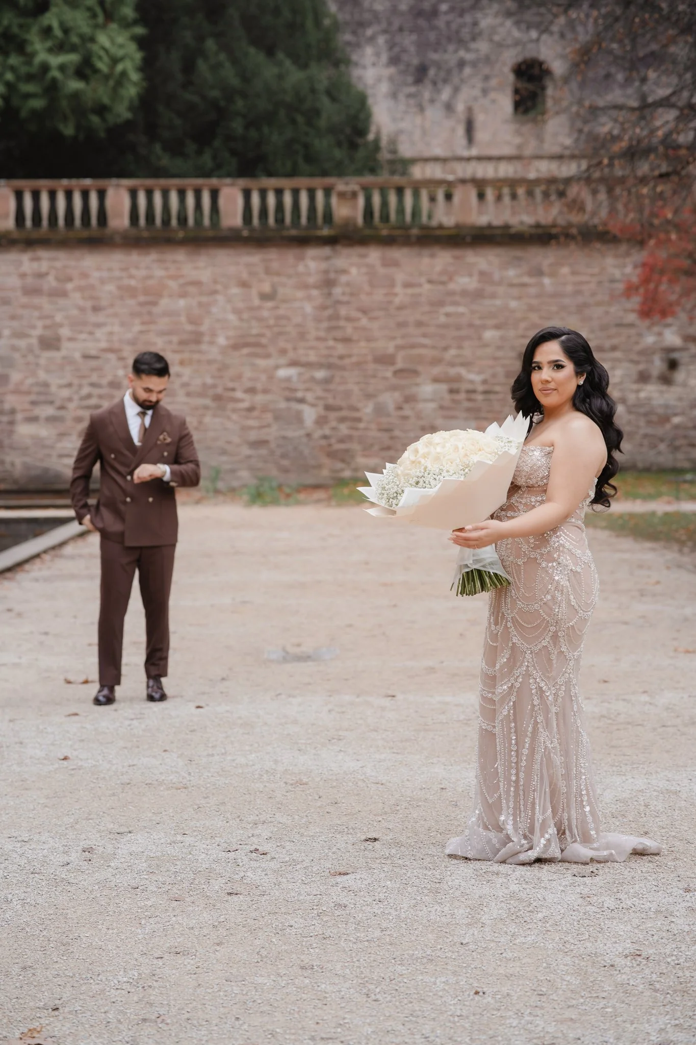 Braut im eleganten beigen Kleid mit Blumenstrauß, Bräutigam im Hintergrund vor Backsteinmauer – Hochzeitsfotografie & Videografie Köln