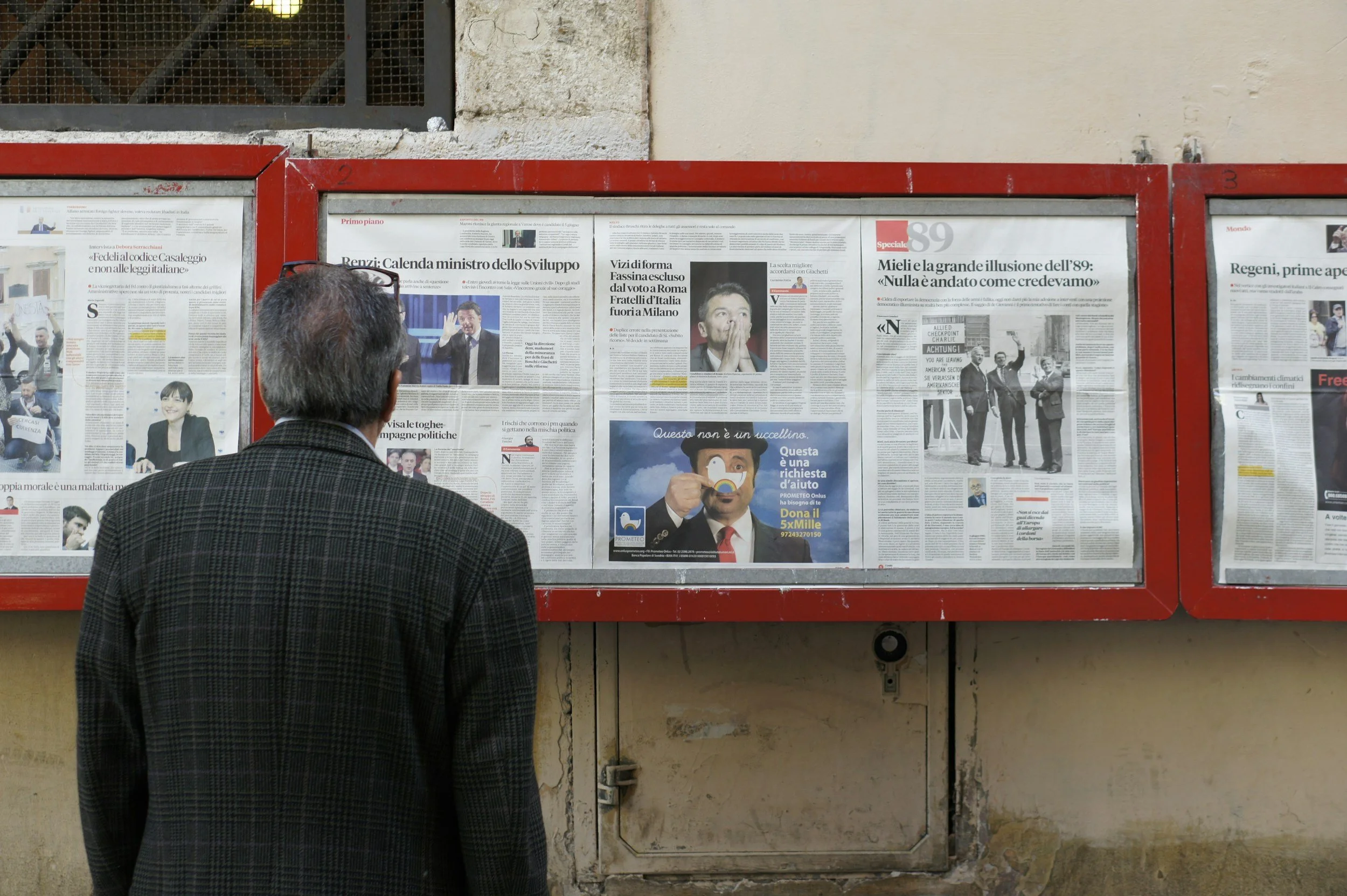 A man with gray hair and glasses on his head is reading newspapers posted on a red bulletin board on a beige wall. The man is wearing a dark checkered jacket.