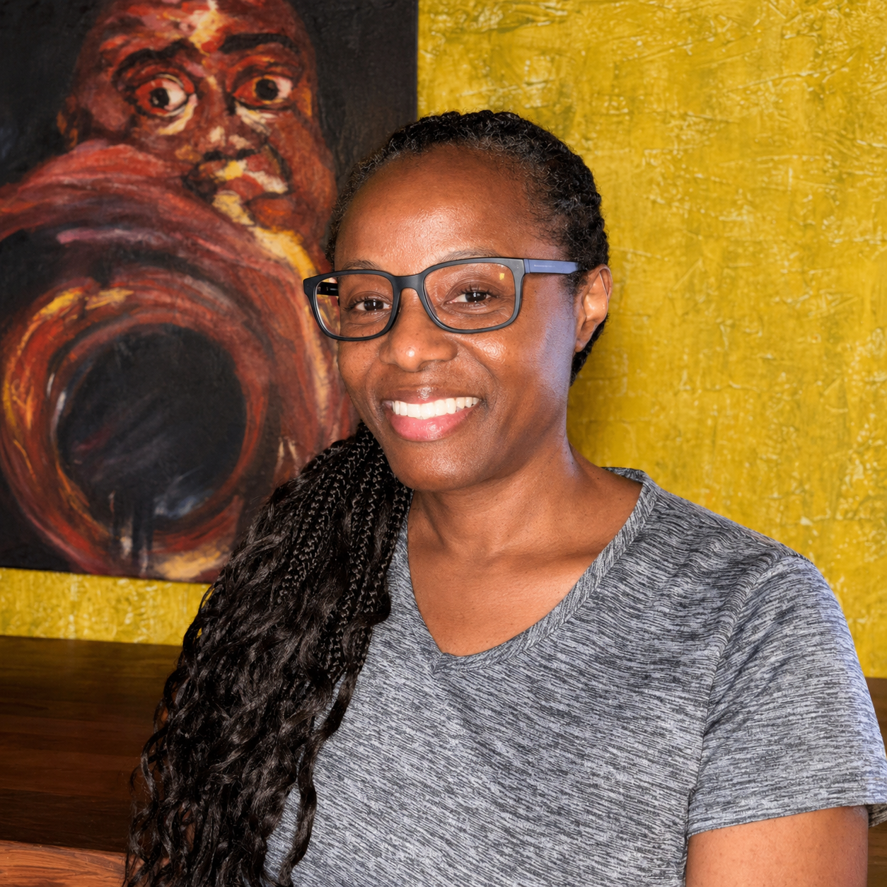A smiling woman with glasses andbraids, wearing a gray shirt, sitting in front of a colorful abstract painting with yellow and dark tones.