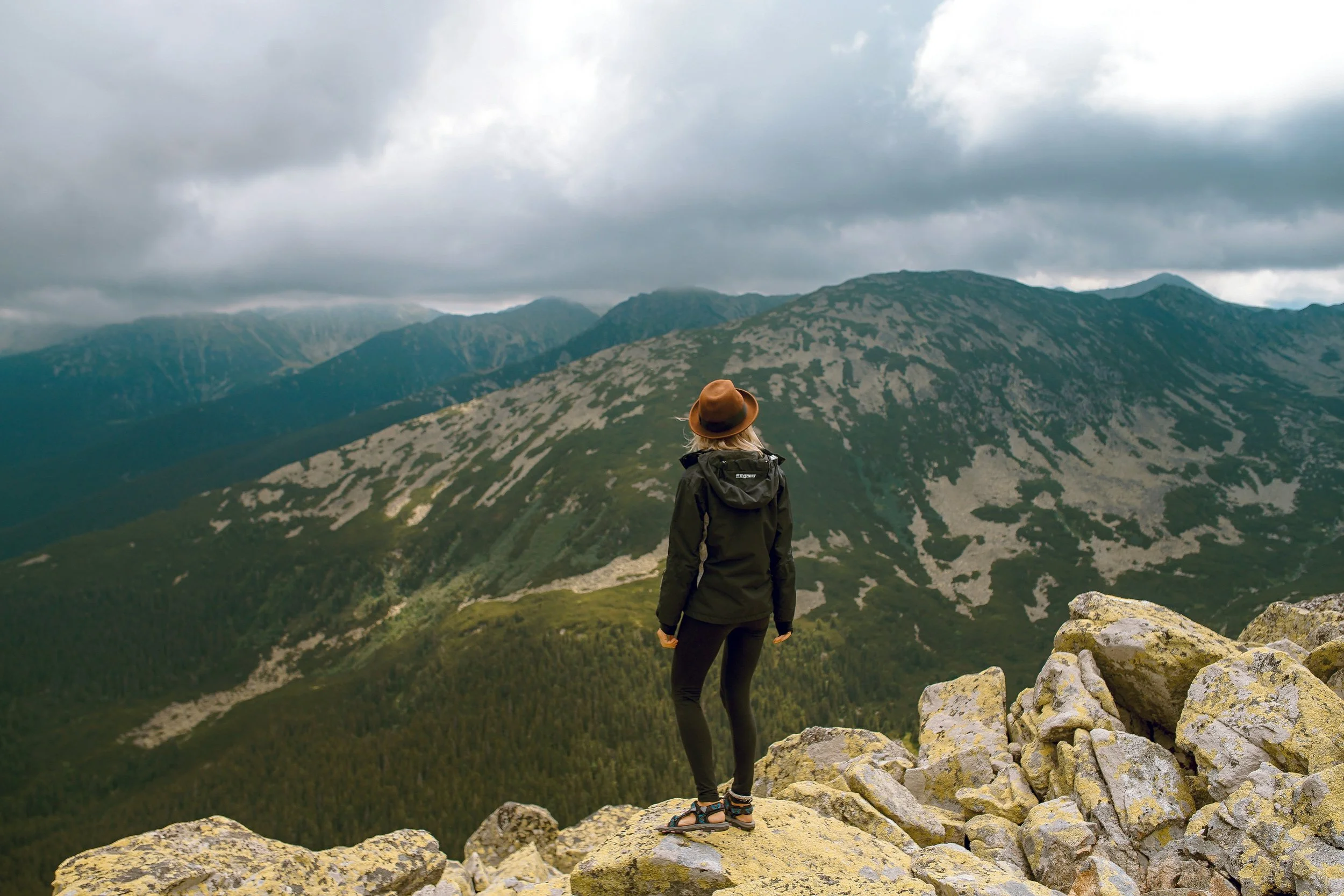 A person with blonde hair, wearing a brown hat and hiking gear, standing on rocks overlooking a mountainous landscape with cloudy skies.