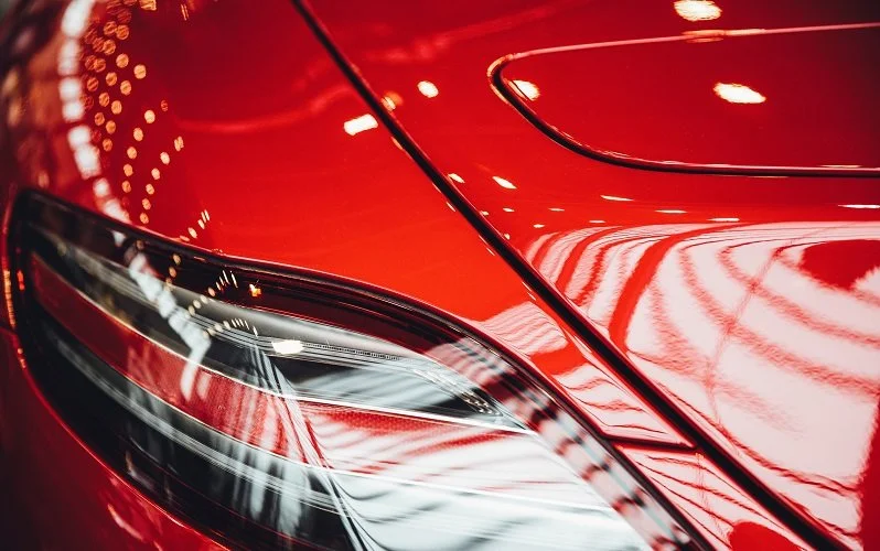 Close-up of the front left corner of a red car, showing the headlight and part of the hood with reflections of indoor lighting.