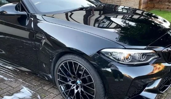 Close-up of a black BMW sports car parked on a brick driveway, showing the front left side, wheel, and part of the hood and windshield with reflections of trees and sky.