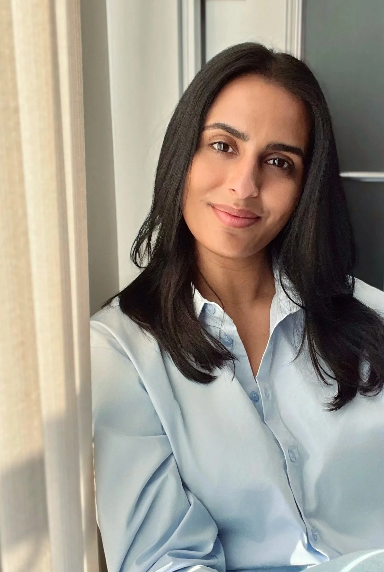 Young woman with shoulder-length dark hair wearing a light blue button-up shirt, smiling, standing near a window.