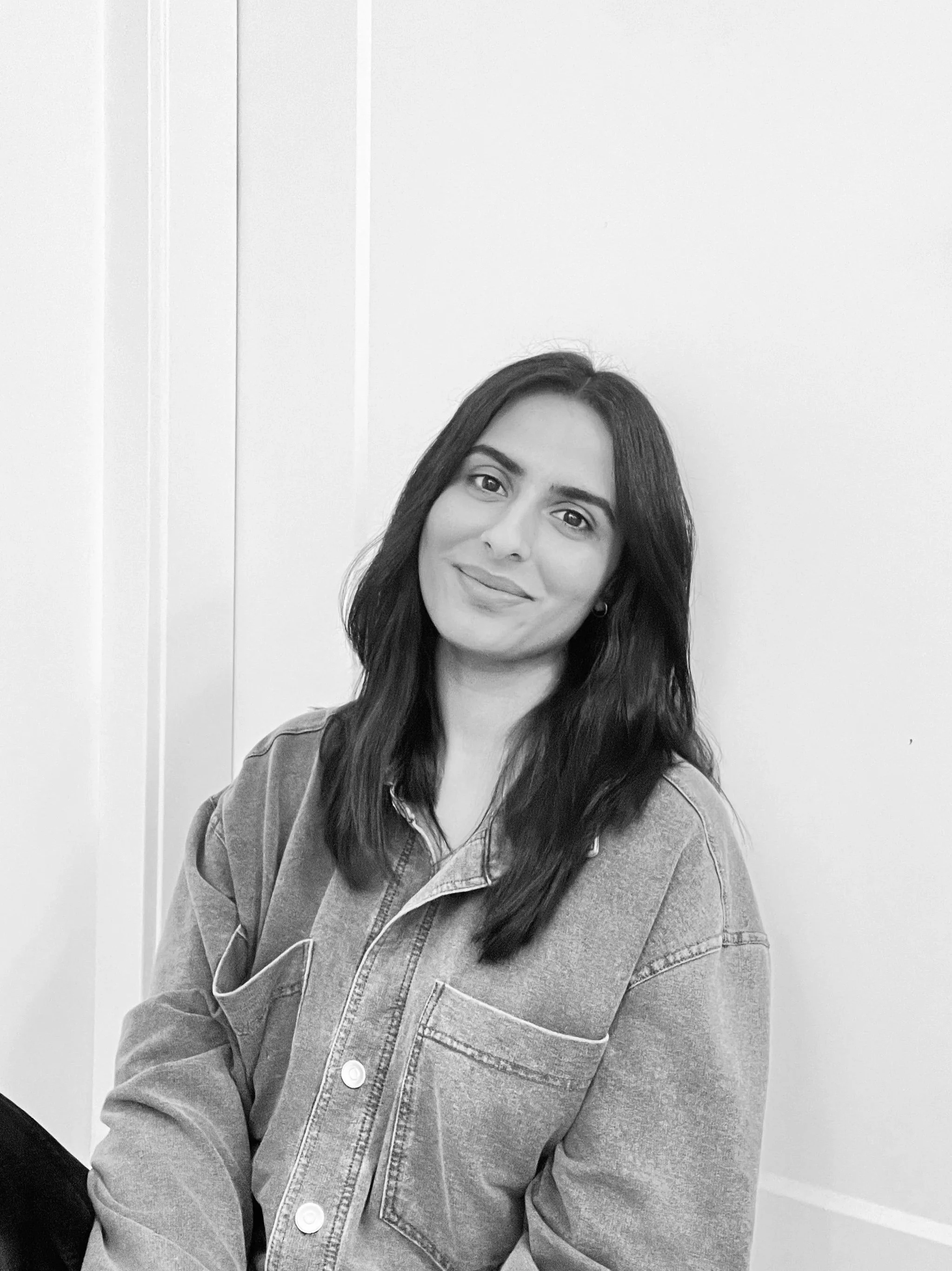 Black and white photo of a young woman with shoulder-length dark hair, wearing a casual denim jacket, smiling softly, standing against a plain wall.