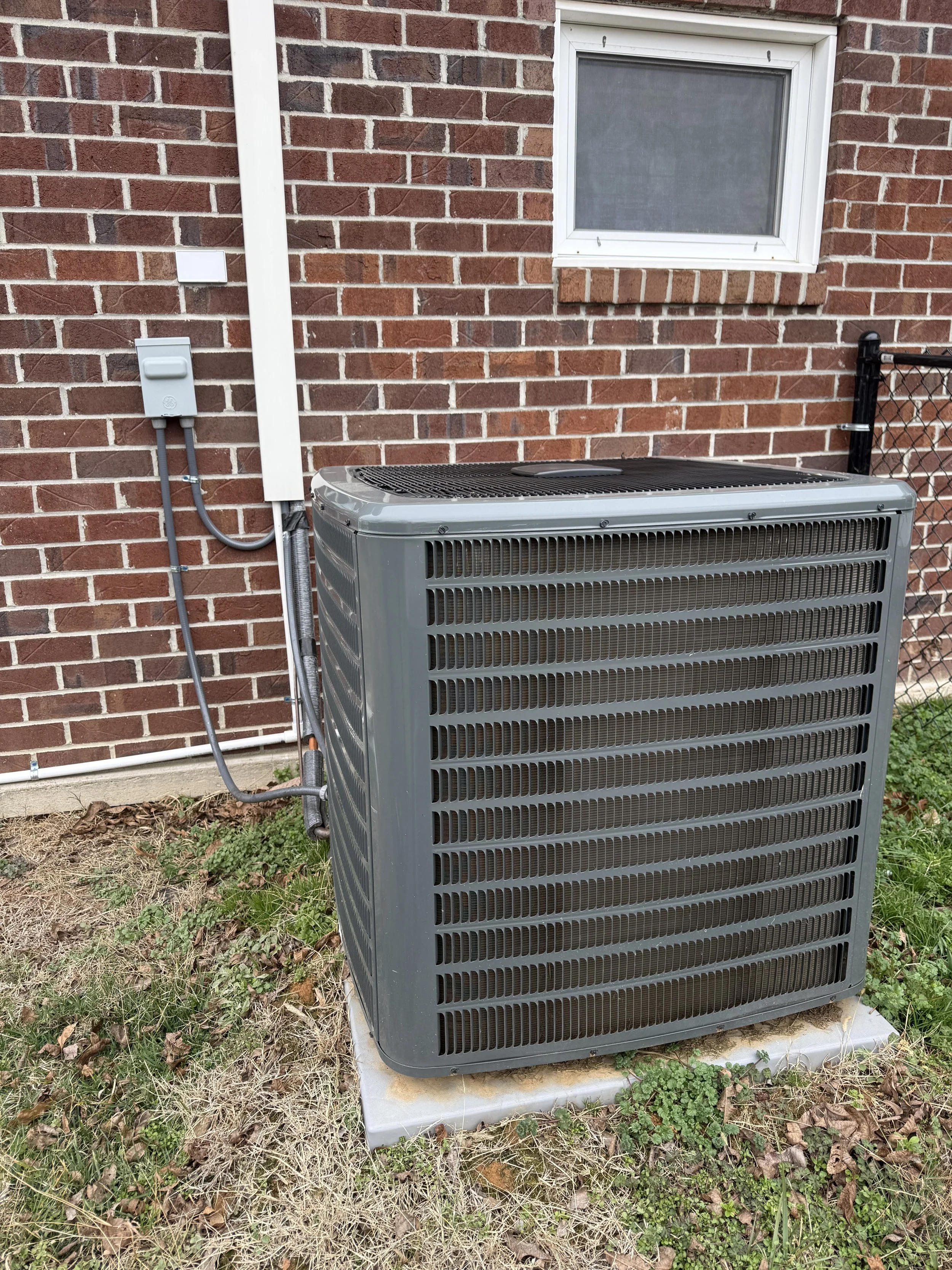 Outside view of a gray central air conditioning unit next to a brick house wall with a window and electrical box.