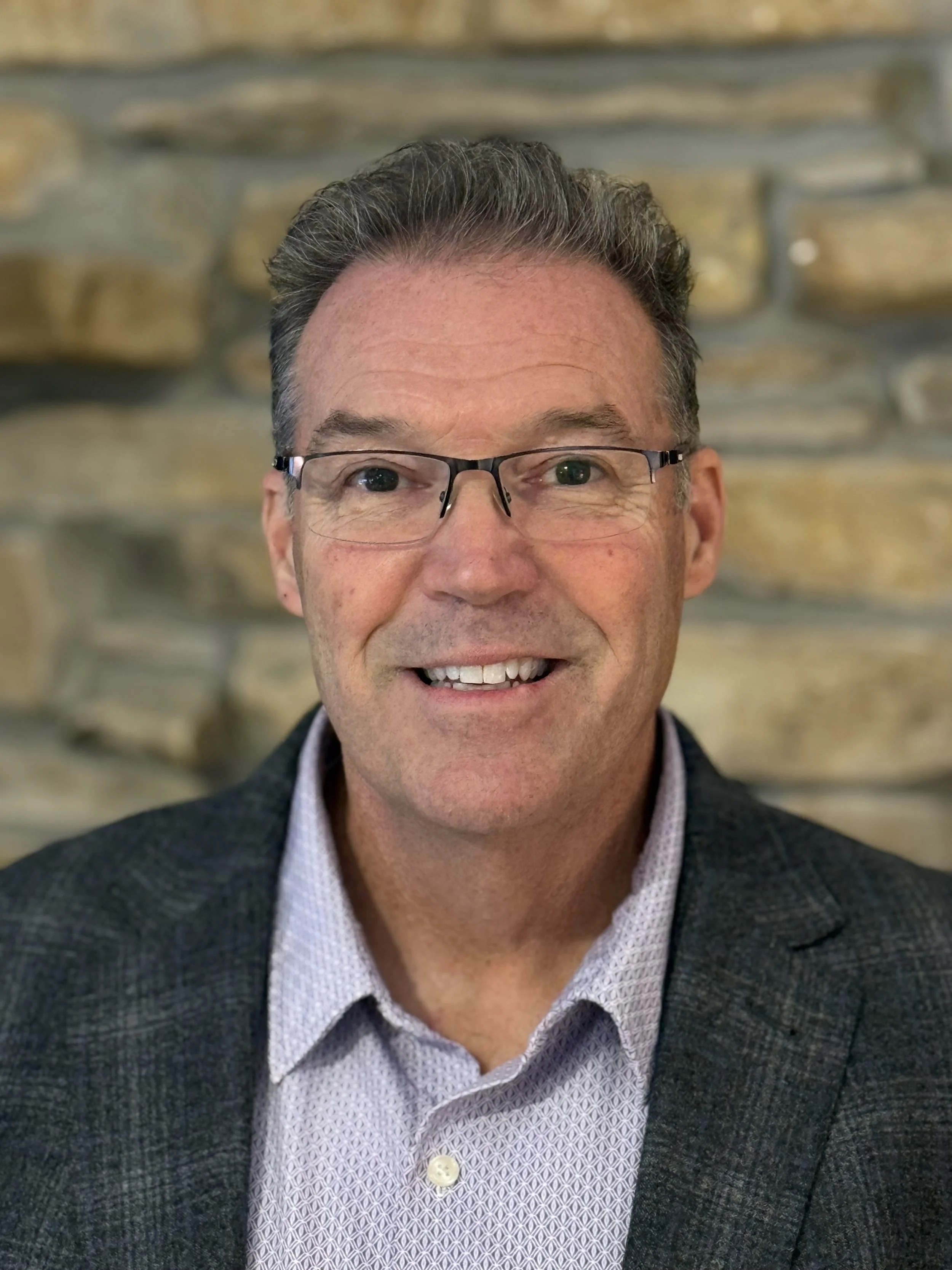 A smiling middle-aged man with short, gray hair, wearing glasses, a light-patterned dress shirt, and a dark blazer, standing in front of a stone wall.