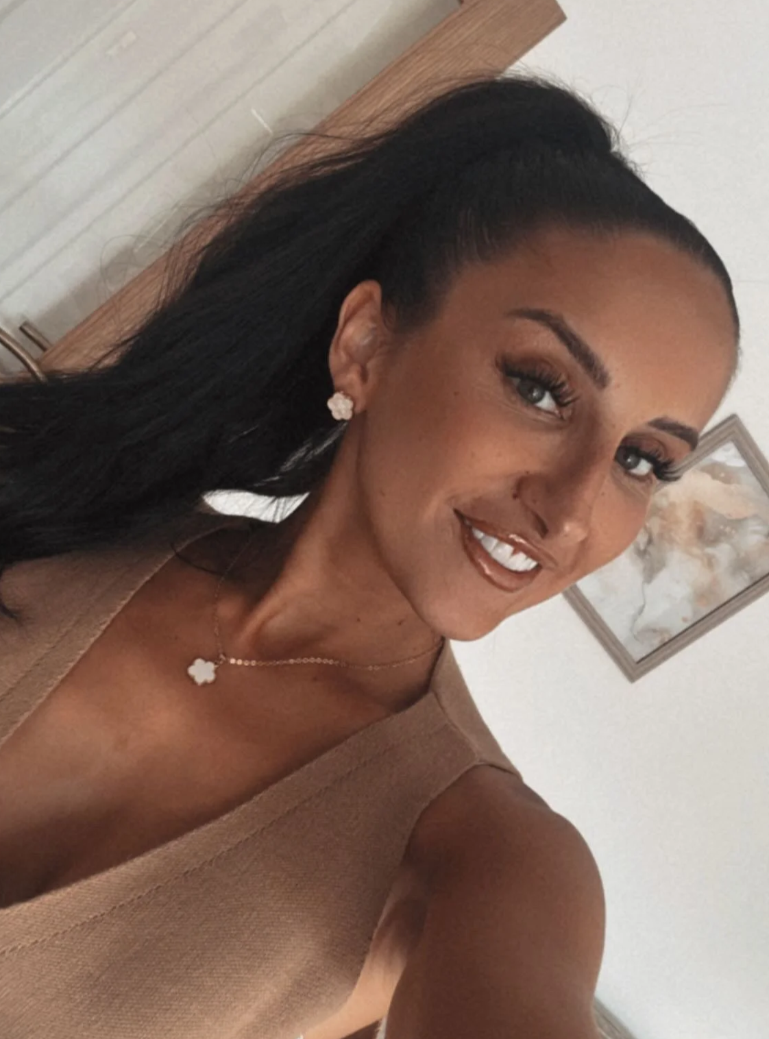 A woman with dark hair styled in a high ponytail, wearing flower-shaped earrings, a necklace with a flower pendant, and a beige sleeveless top, smiling at the camera.