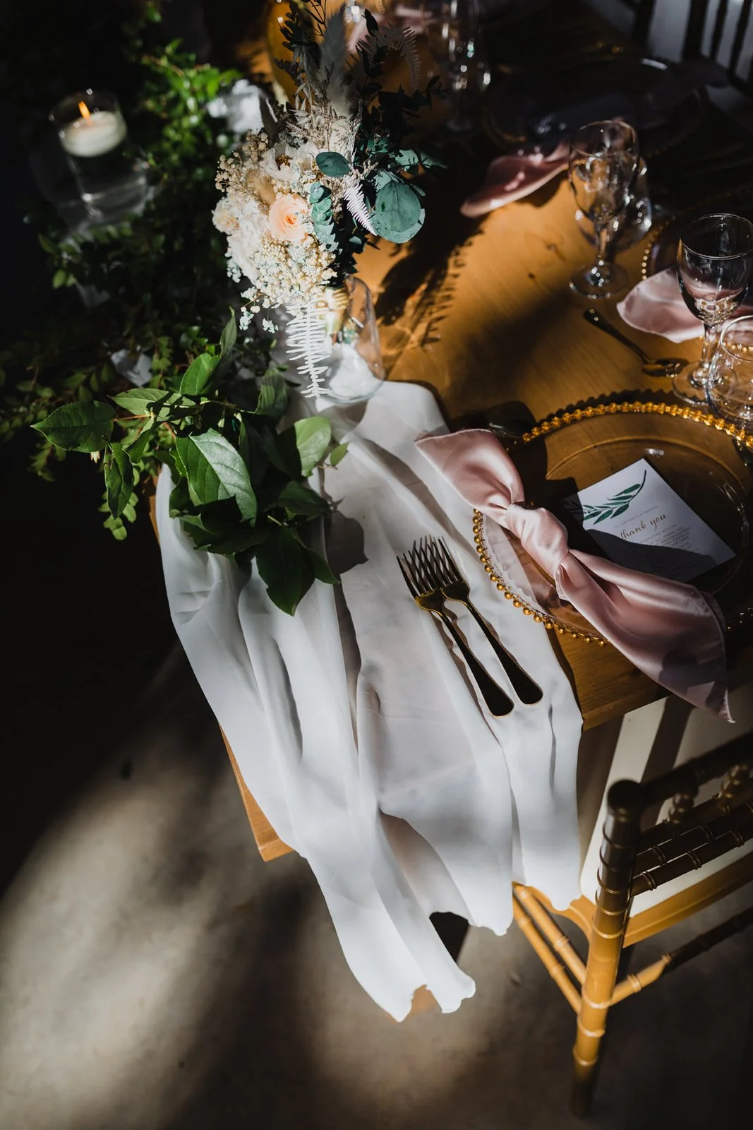 Elegant table setting with a floral centerpiece, pink satin ribbon on a glass plate, gold cutlery, wine glasses, and a white table runner.
