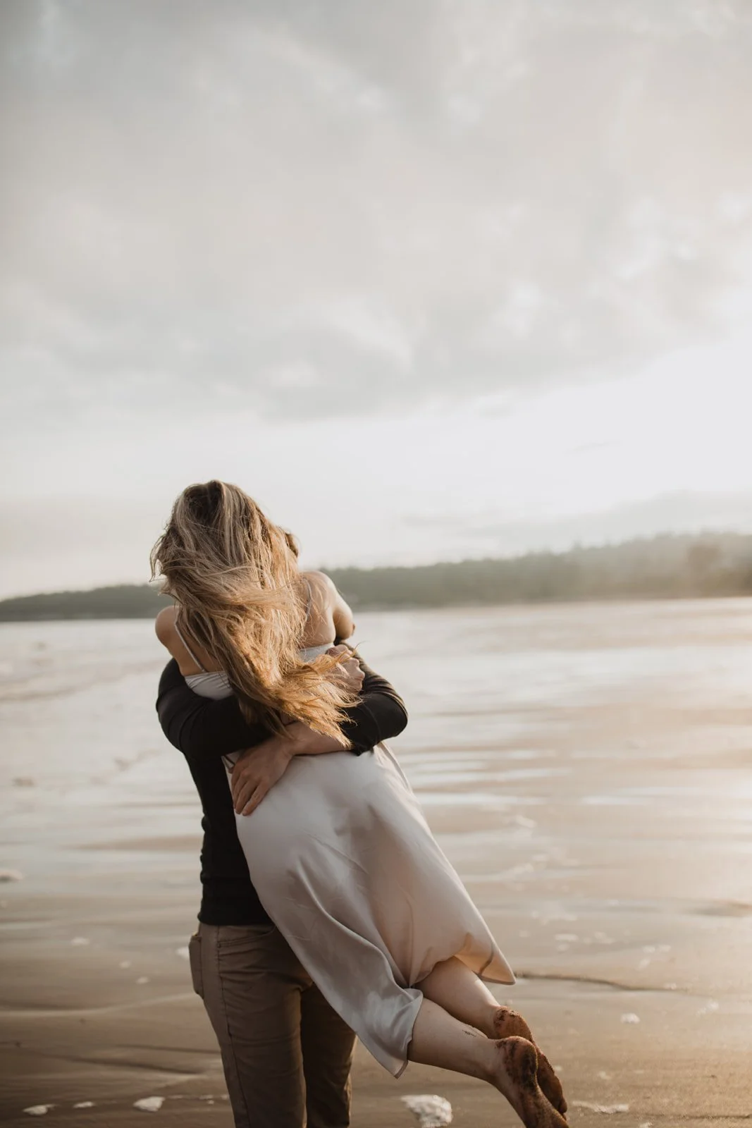 Person holding a woman in their arms on a beach with cloudy sky in the background.
