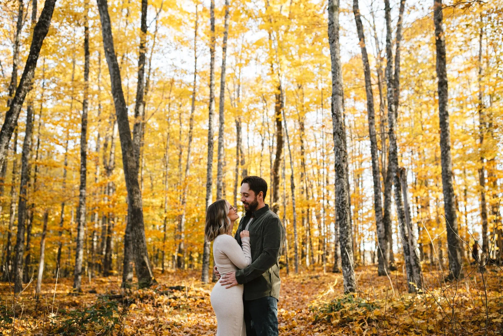 A couple standing close together in a forest during fall, surrounded by yellow and orange leaves on trees and ground.