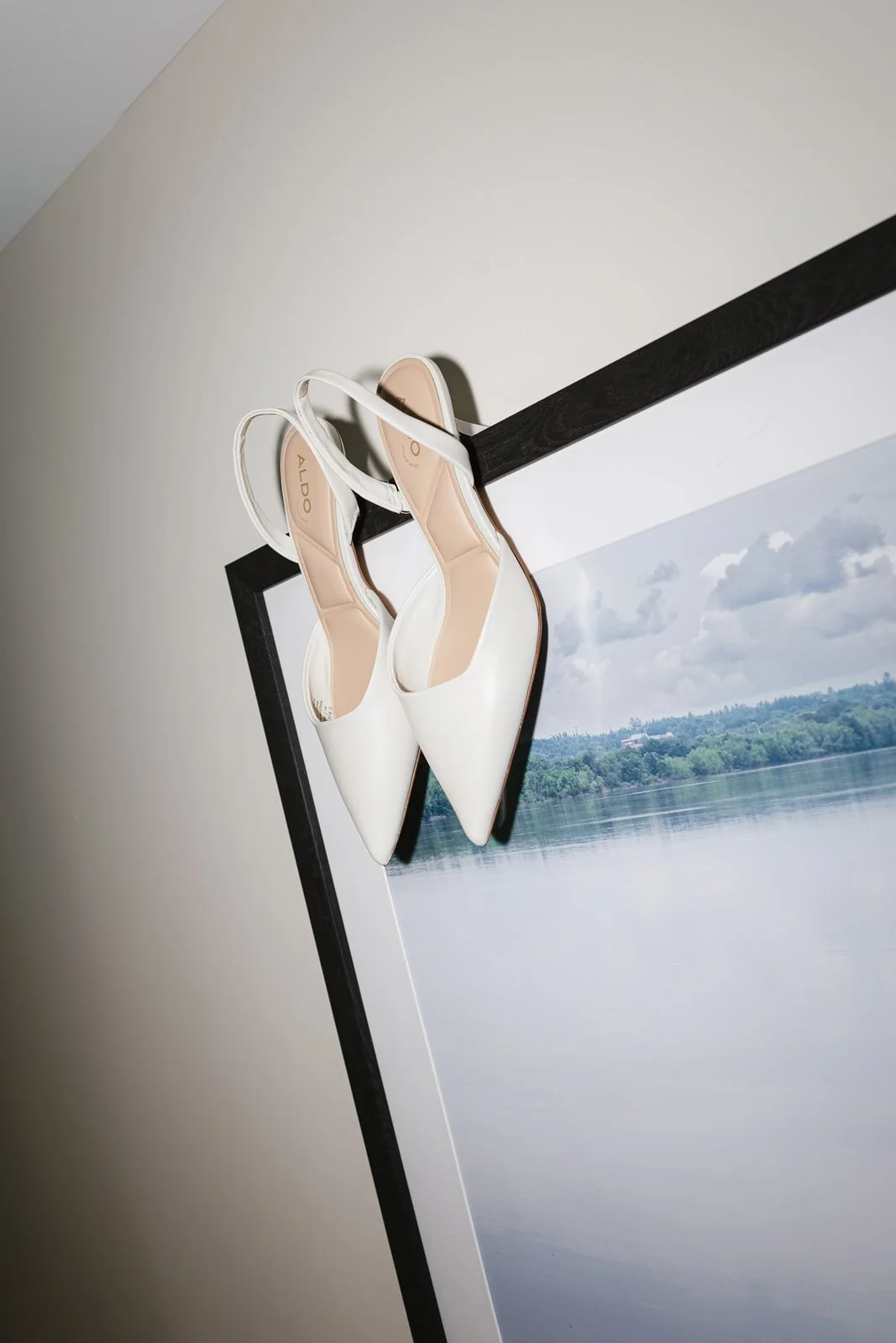 White high-heeled shoes hanging on the edge of a picture frame leaning against a white wall.