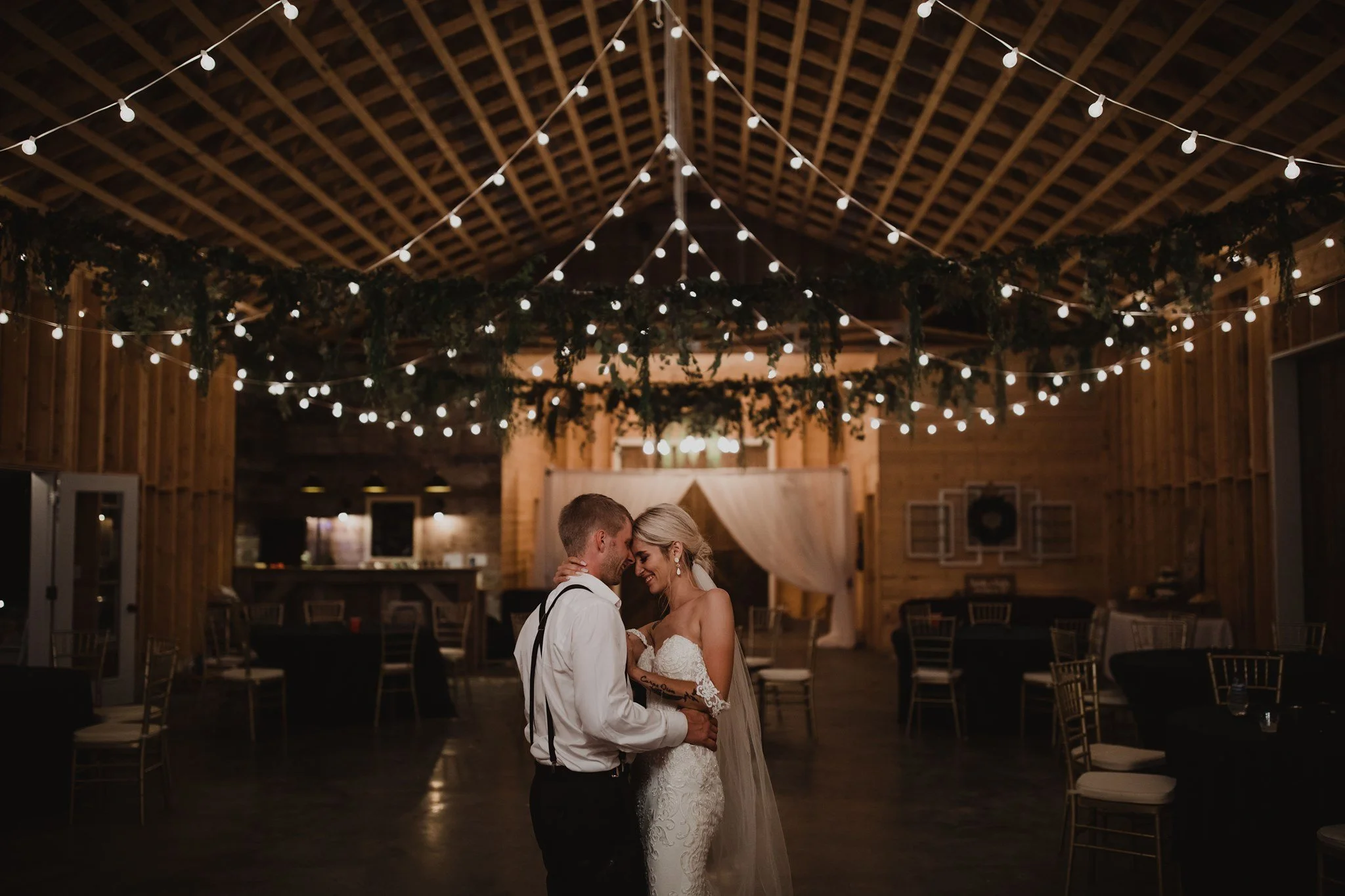 A bride and groom share a dance in a rustic barn decorated with string lights and greenery at their wedding reception.