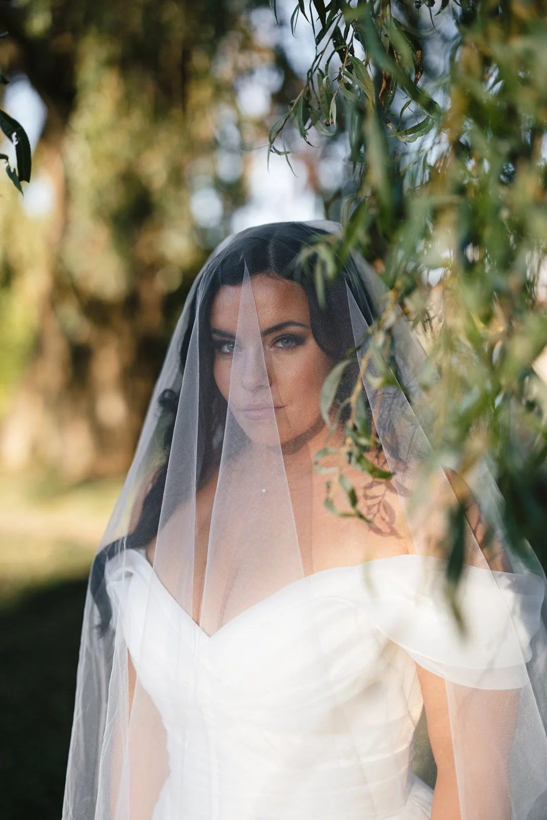 A bride with dark hair and blue eyes, wearing a white wedding dress and veil, partially obscured by green foliage outdoors.
