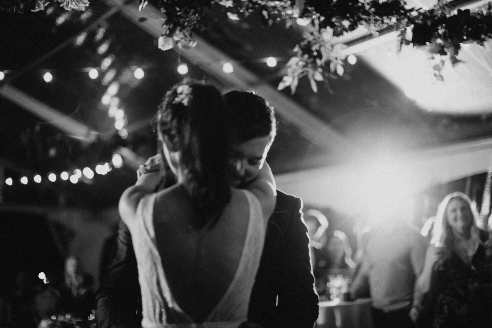 A couple dancing closely at a reception, the man in a tuxedo and the woman in a sleeveless gown, with string lights and people in the background.