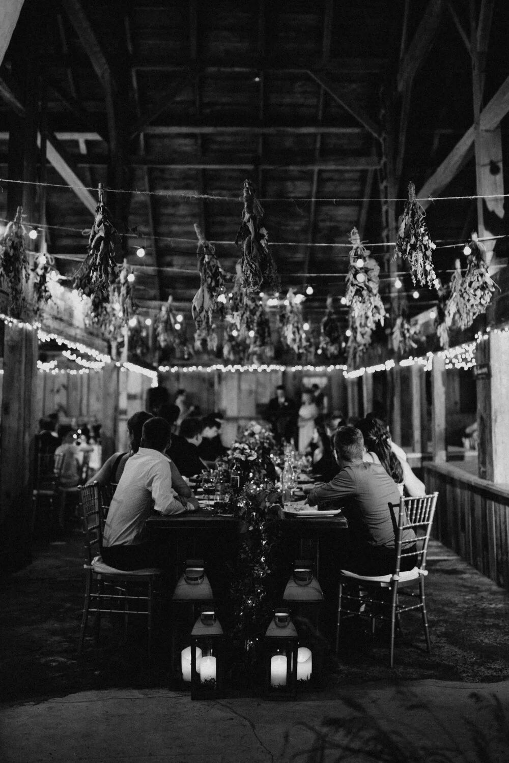 Guests seated at a long table decorated with flowers and lanterns, inside a rustic barn with hanging dried herbs and string lights, during a nighttime event.
