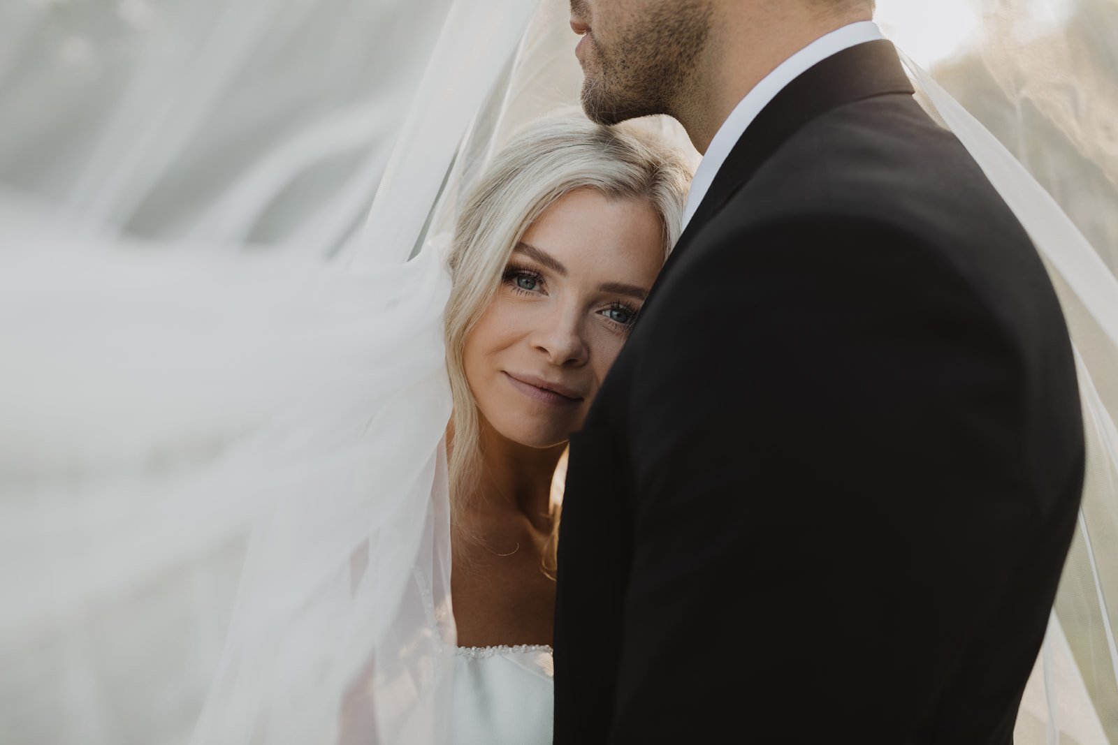 A bride with blonde hair and blue eyes looking at the camera, partially obscured by her wedding veil, and a groom in a black tuxedo standing close to her, with his face turned away, during a wedding photoshoot.