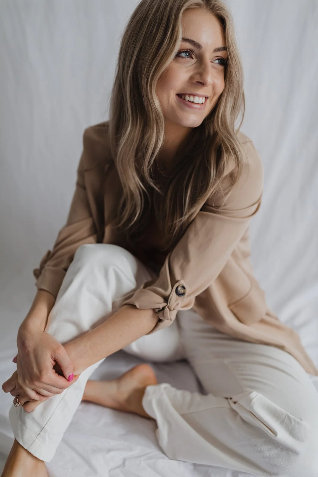 A woman with long wavy blonde hair wearing a beige blazer and white pants, sitting on a bed, smiling and looking to her right.