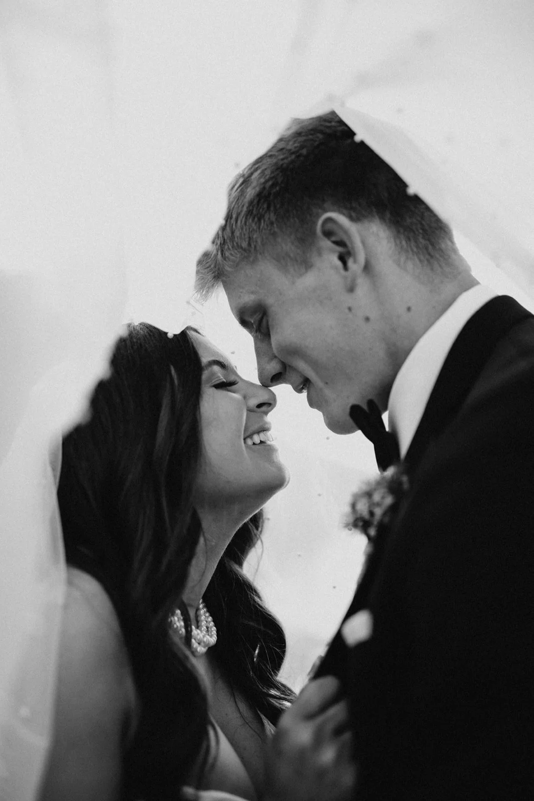 Black and white photo of a bride and groom close together, smiling and touching foreheads, with the bride wearing a pearl necklace and the groom in a tuxedo and bow tie.