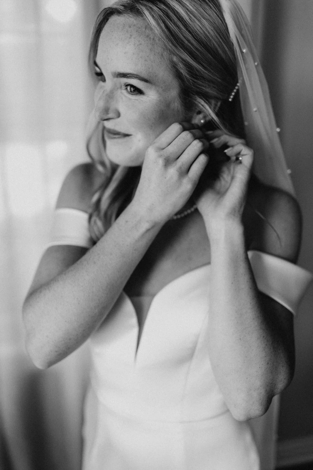A black-and-white photograph of a young bride adjusting her earrings, wearing a wedding dress and veil.