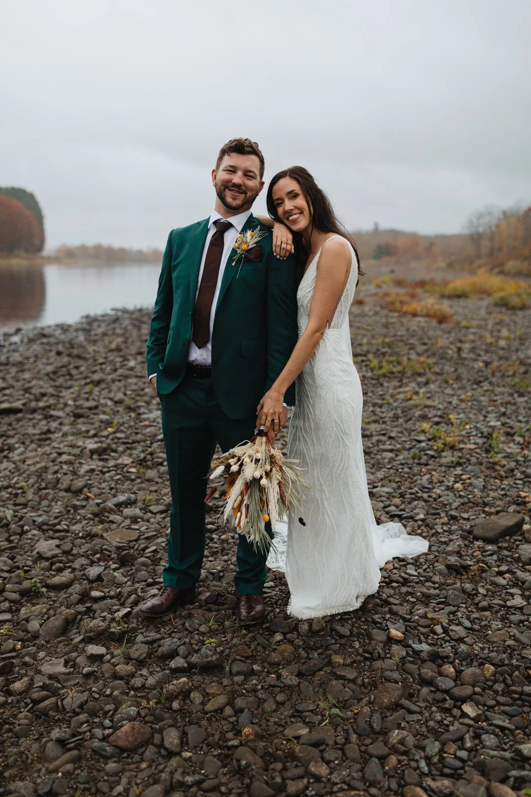 A newlywed couple standing on a rocky shoreline near a lake, smiling at the camera. The groom wears a green suit with a white shirt and brown tie, holding a bouquet of dried flowers. The bride wears a white lace wedding dress, resting her arm on the 