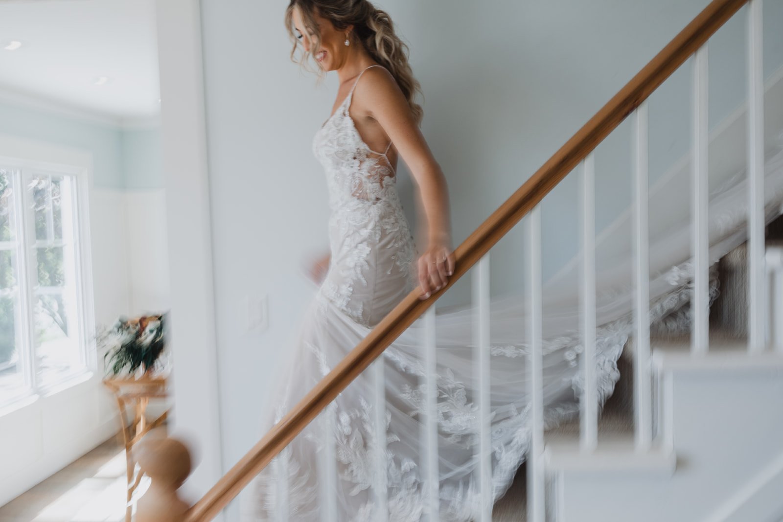 A woman in a white lace wedding dress ascending a staircase in a bright room with large windows