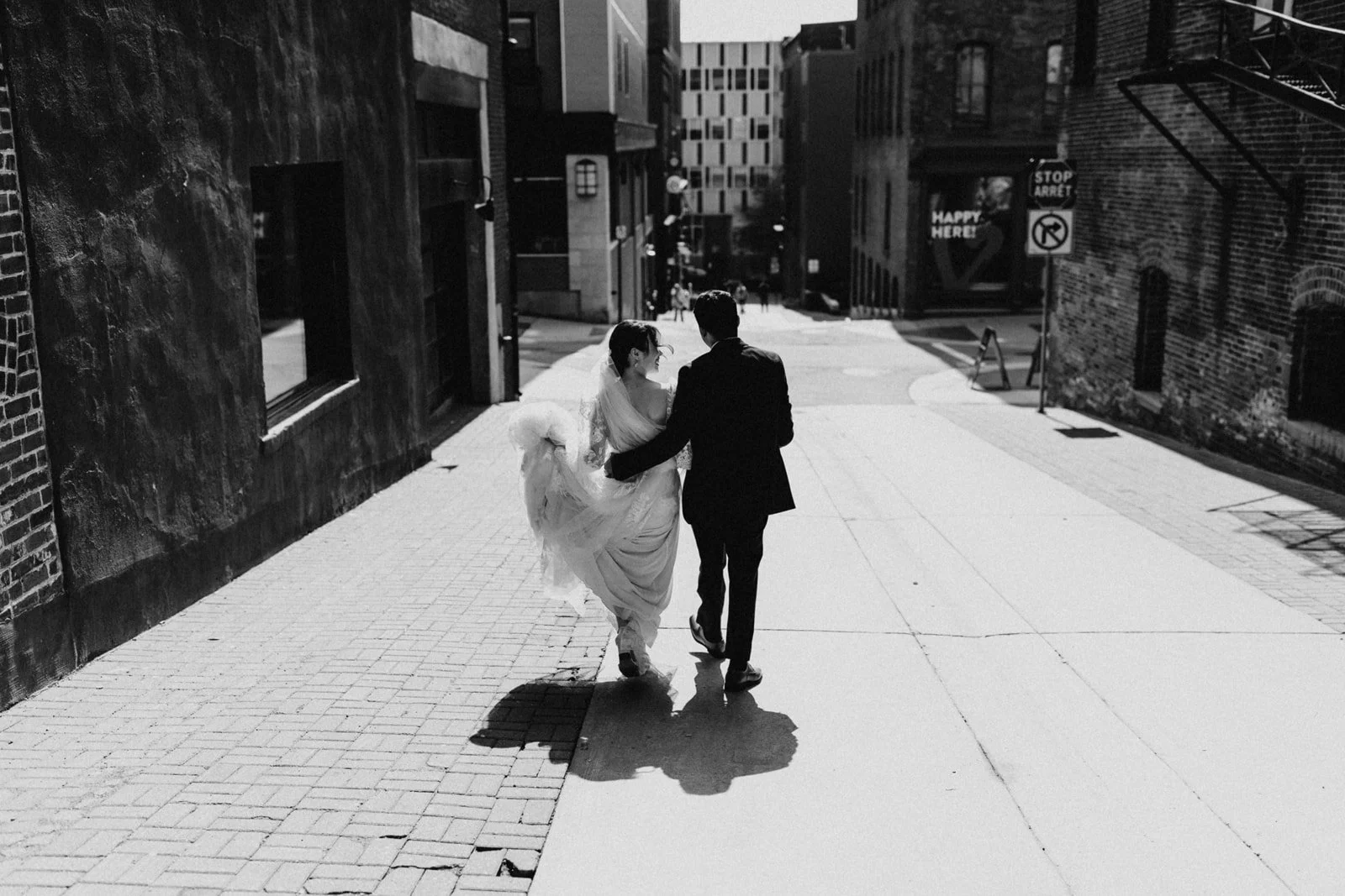 A black and white photo of a couple walking together on a city street, with the woman in a wedding dress and the man in a suit. They are holding hands and appear to be happy.