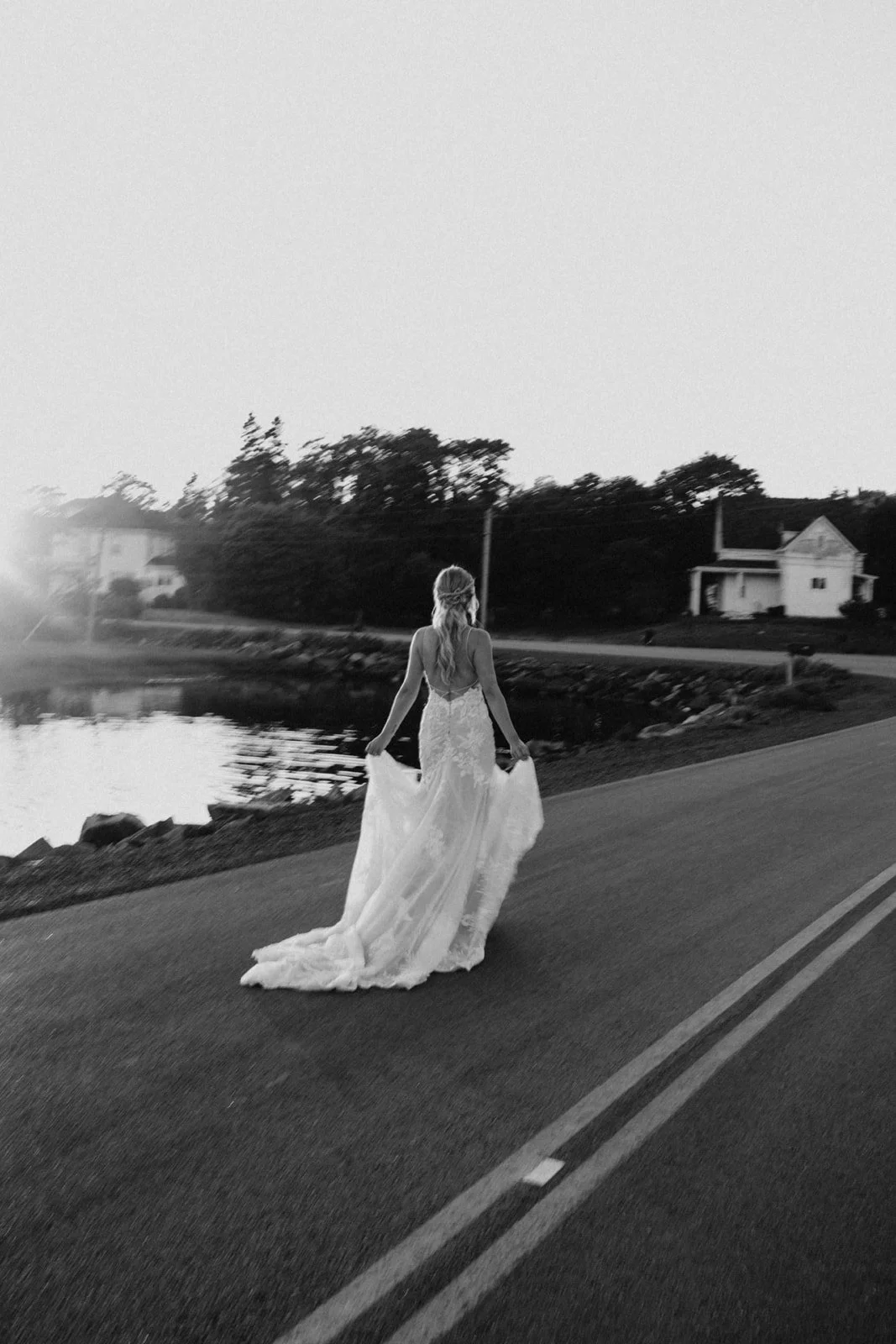 A woman in a wedding dress walking along a road beside a body of water during sunset or sunrise, with trees and houses in the background.
