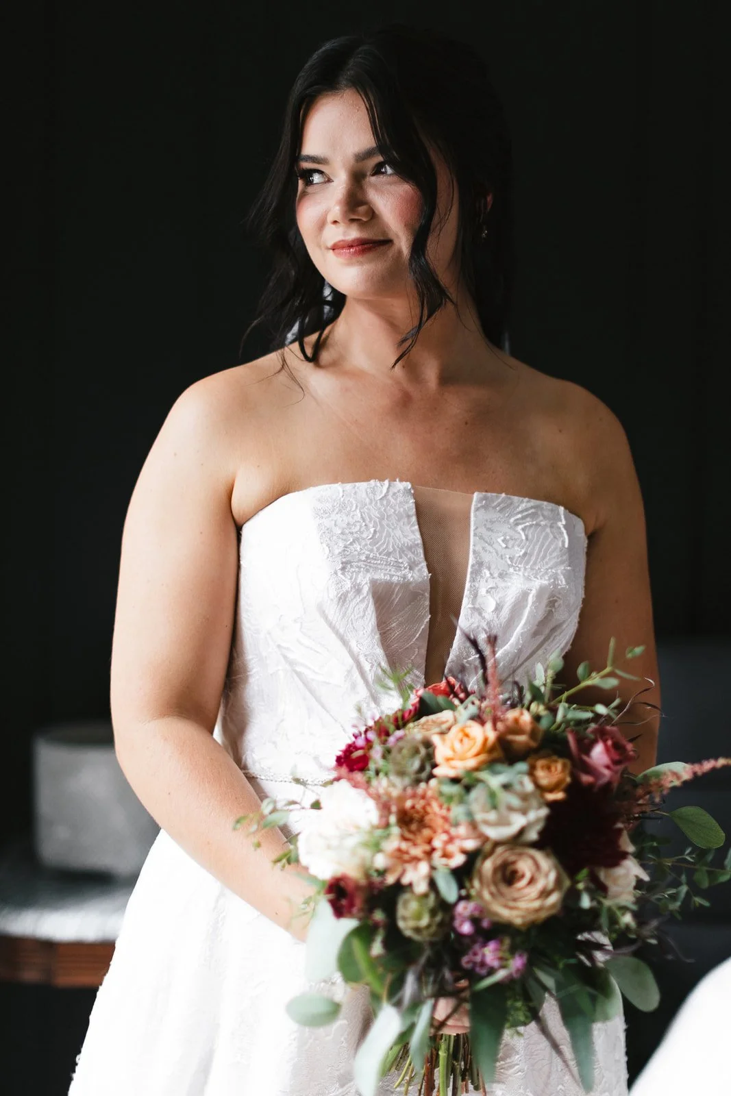 A woman in a white wedding dress holding a bouquet of flowers, standing against a dark background.