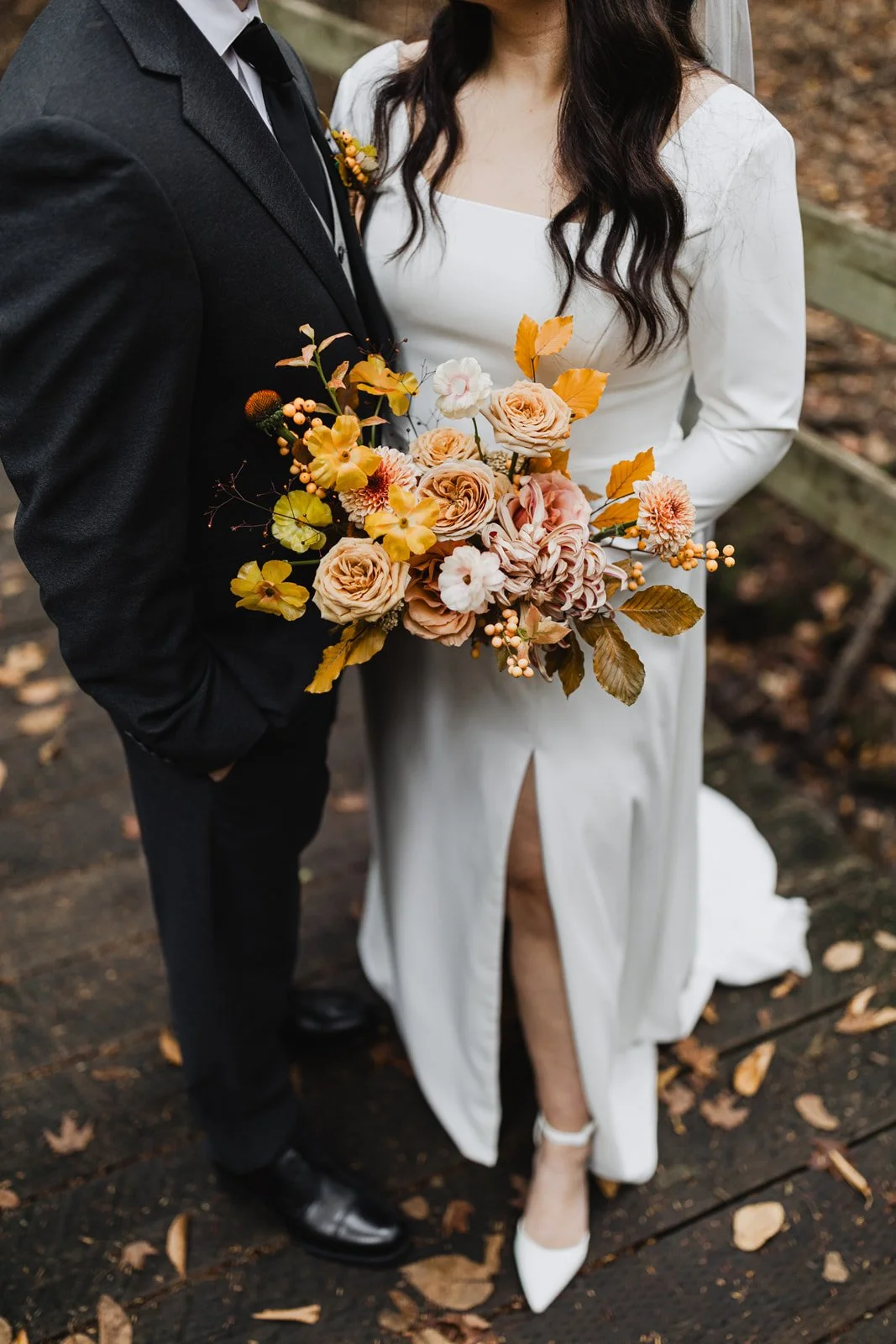 A bride and groom stand on a wooden pathway during a wedding, with the bride holding a bouquet of yellow and peach-colored flowers.