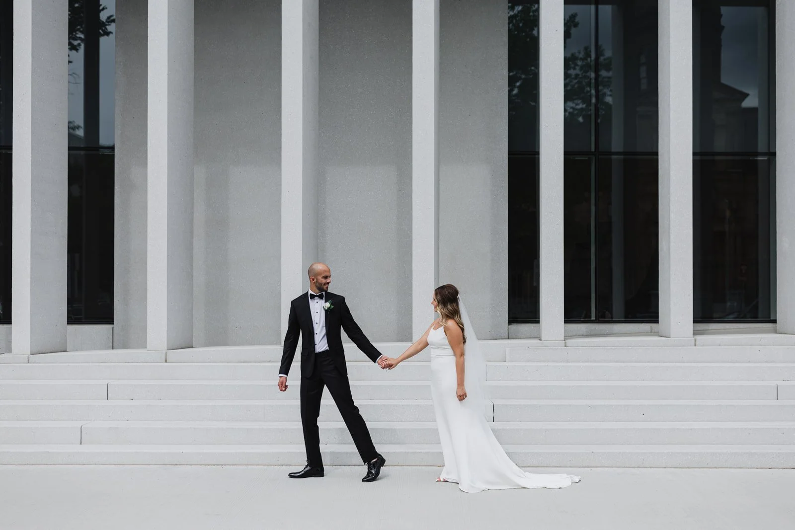 A bride and groom holding hands on the steps of a modern building with large glass windows.