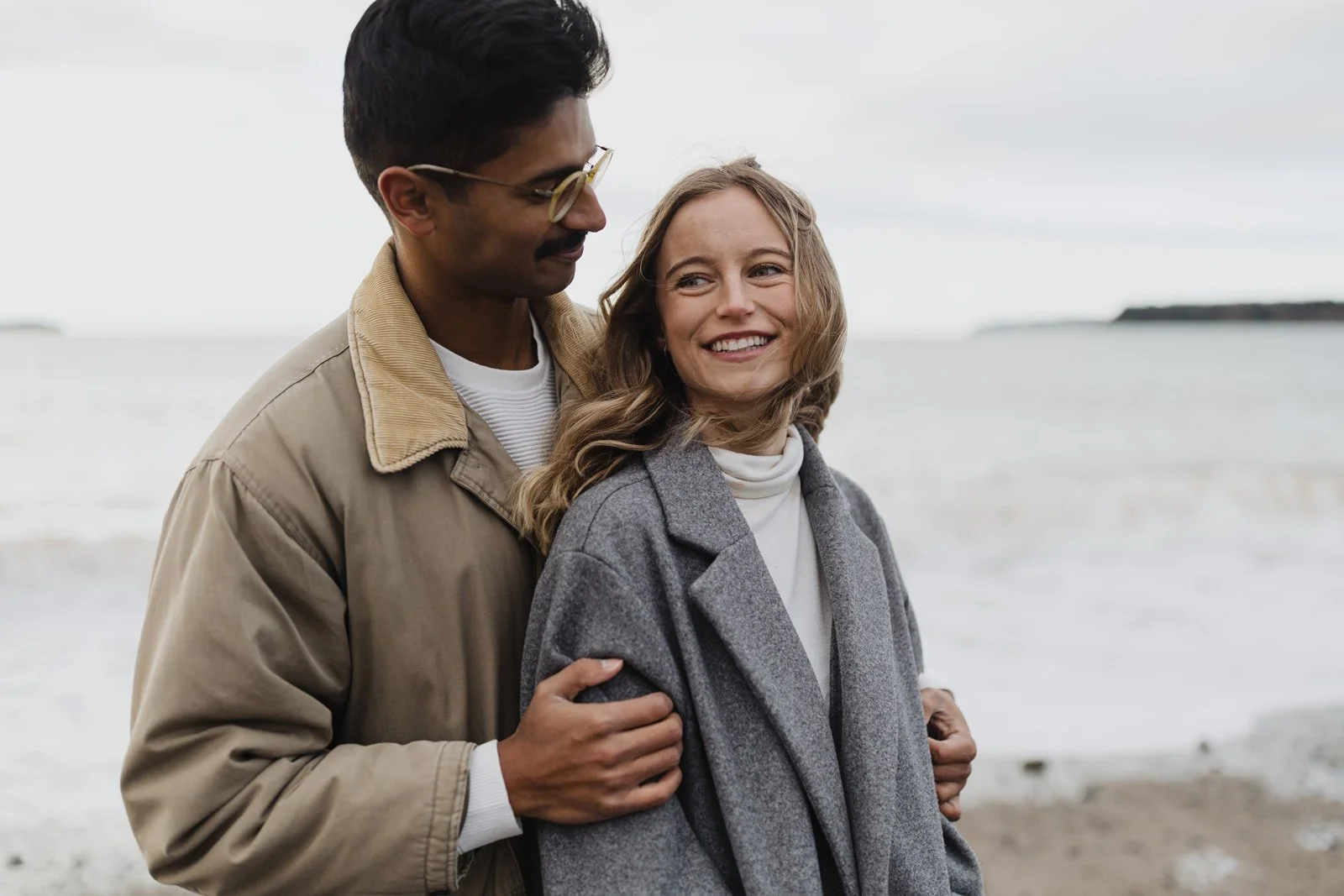 A couple at the beach, with the man holding the woman's arm from behind. They are smiling and looking at each other. Overcast sky with water and distant land in the background.