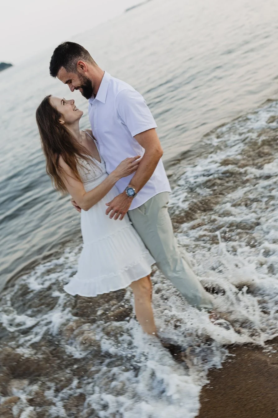 A happy couple stands in shallow water on a beach, embracing and smiling at each other with the ocean in the background.
