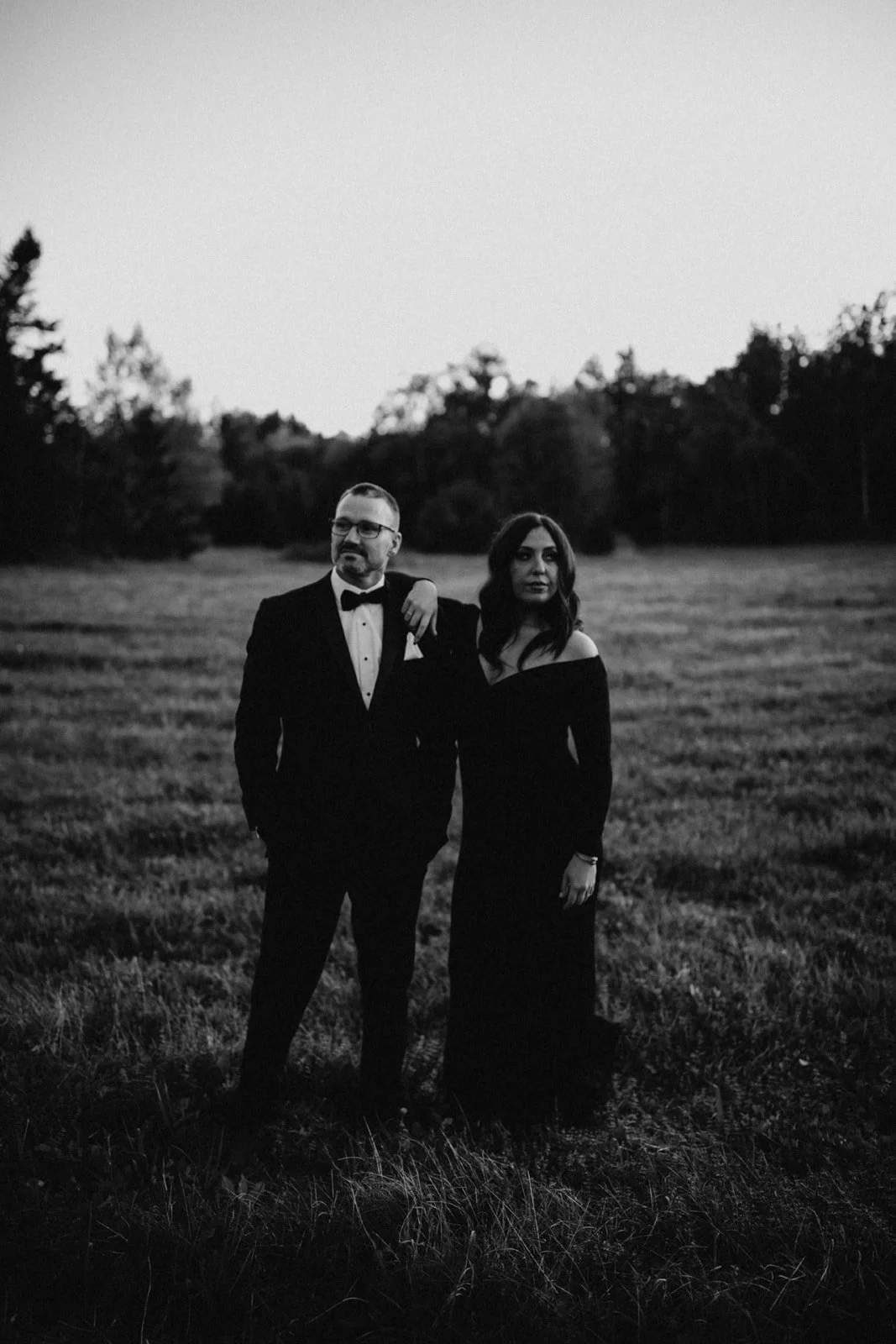 A black and white photo of a man in a tuxedo and a woman in a long black dress standing outdoors in a field at dusk.