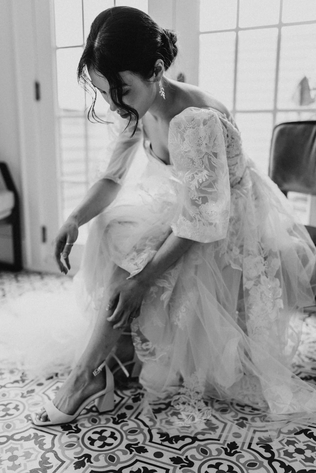 A woman in a wedding dress is putting on her high heels. She is bending down and adjusting her shoe while looking at her leg. The room has patterned tile flooring and furniture in the background.