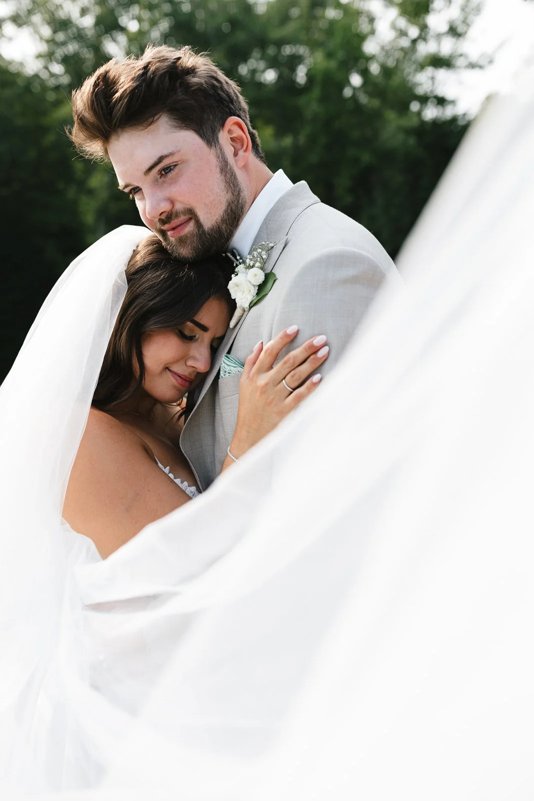 A bride and groom embrace outdoors during their wedding day, with the bride resting her head on the groom's chest and the groom looking content, both surrounded by greenery and daylight.