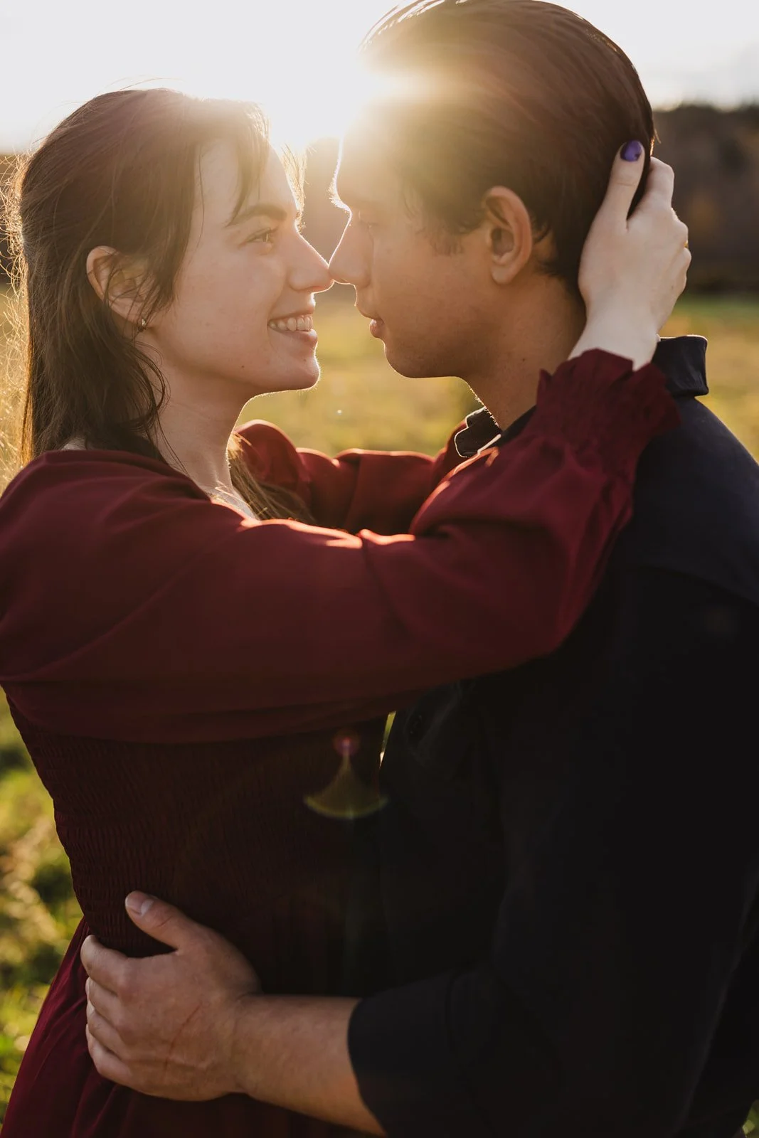 A young couple embraces outdoors at sunset, facing each other closely, with sunlight shining behind them.