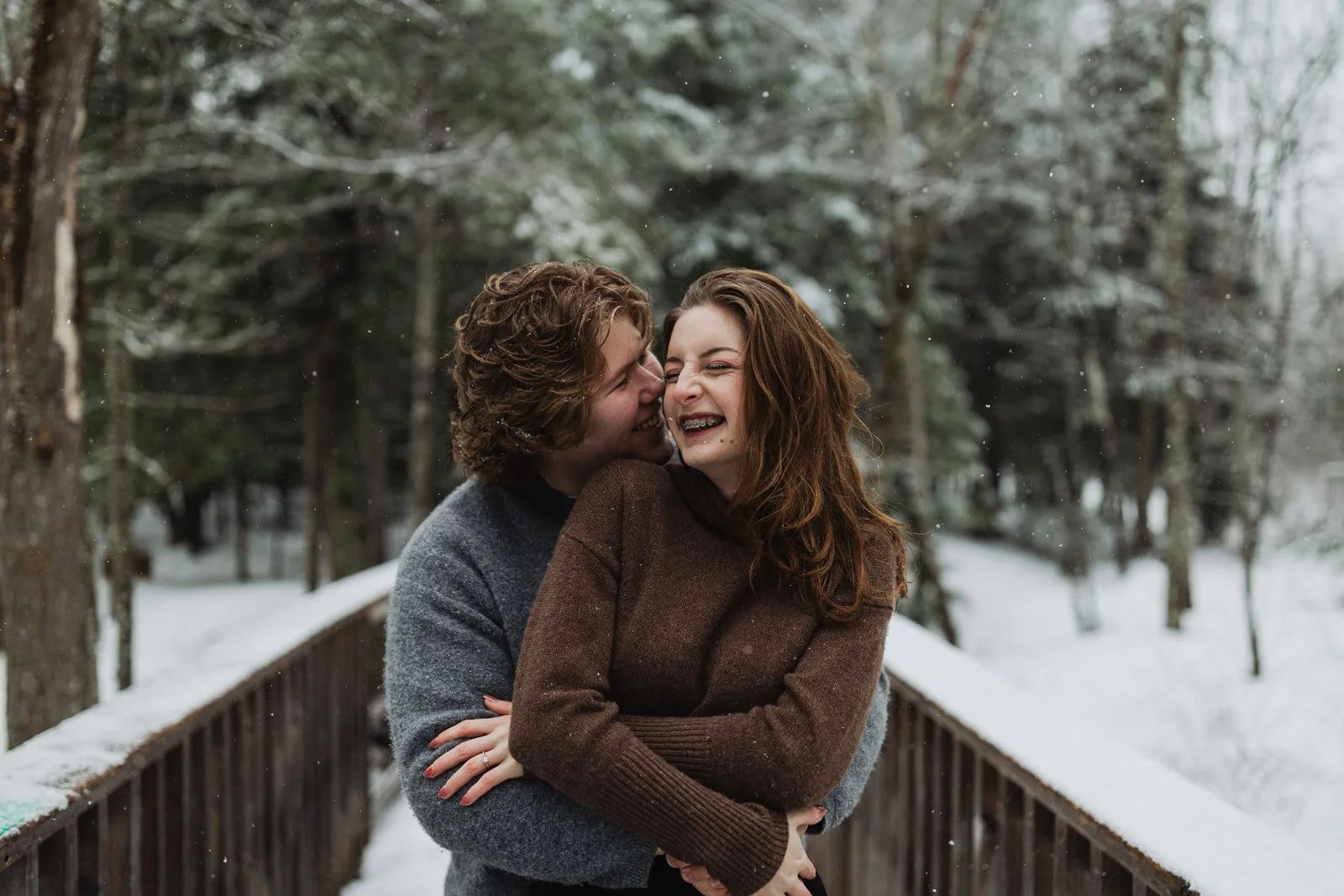 A couple laughing and embracing on a snow-covered bridge in a winter forest.